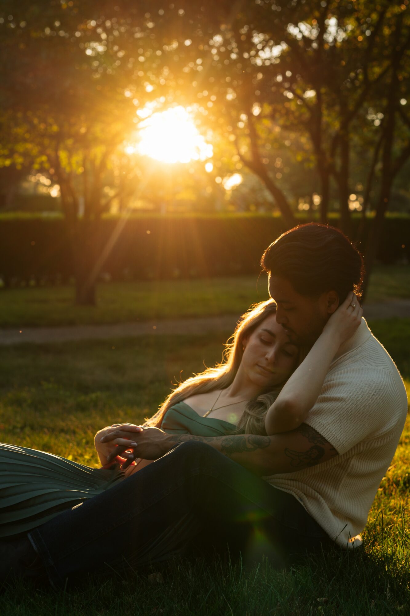 A couple sitting on grass in a park during sunset, embracing each other with trees and sunlight in the background.