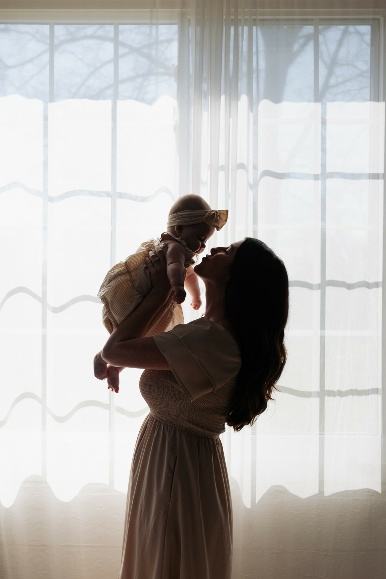 Silhouette of a woman holding a child in front of a window with curtains, backlit by daylight.