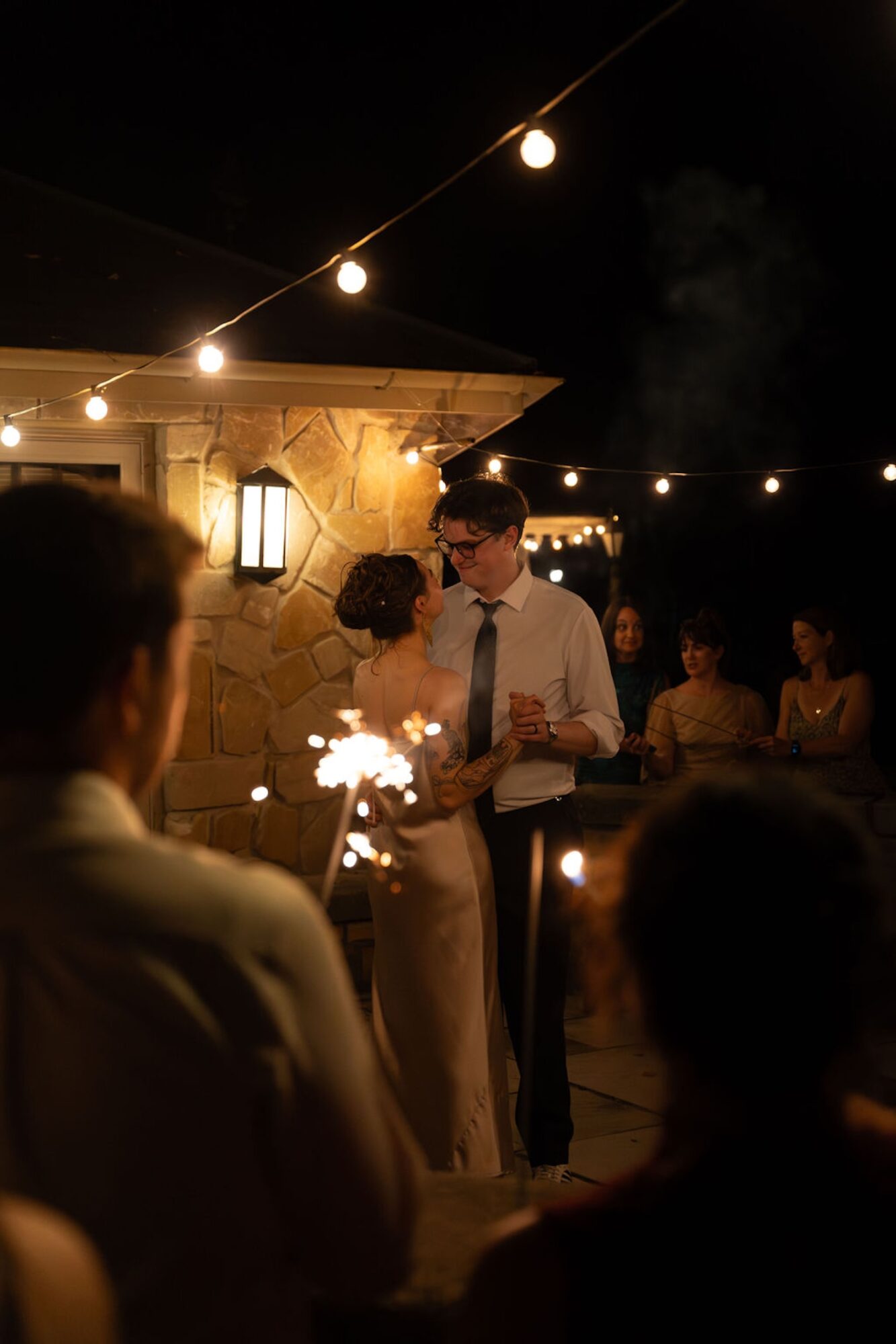 People gather outdoors at night under string lights, with a couple dancing and sparklers, illuminated by warm lighting.