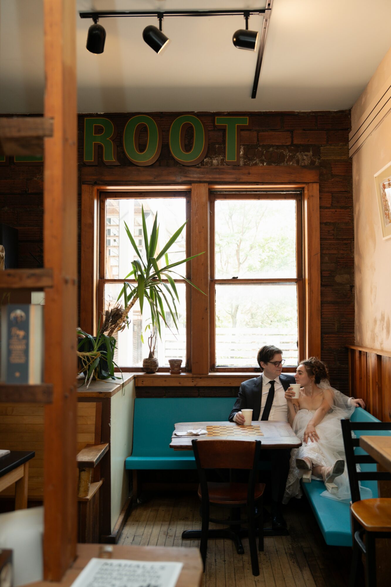 People sitting at a table in a cozy cafe with a large window, plants, and a sign reading 'ROOT' above the window.