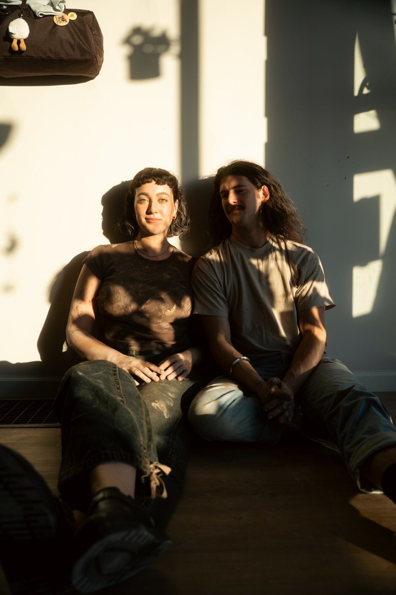 Two women sit on a bench with sunlight casting shadows on the wall behind them, one with curly hair and the other with straight hair.