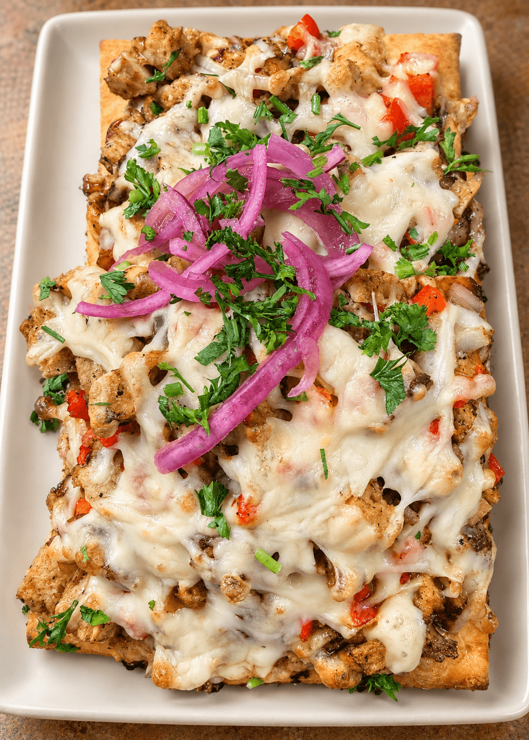 Baked dish with melted cheese, chopped vegetables, and pickled red onions on top, garnished with herbs on a rectangular white plate.