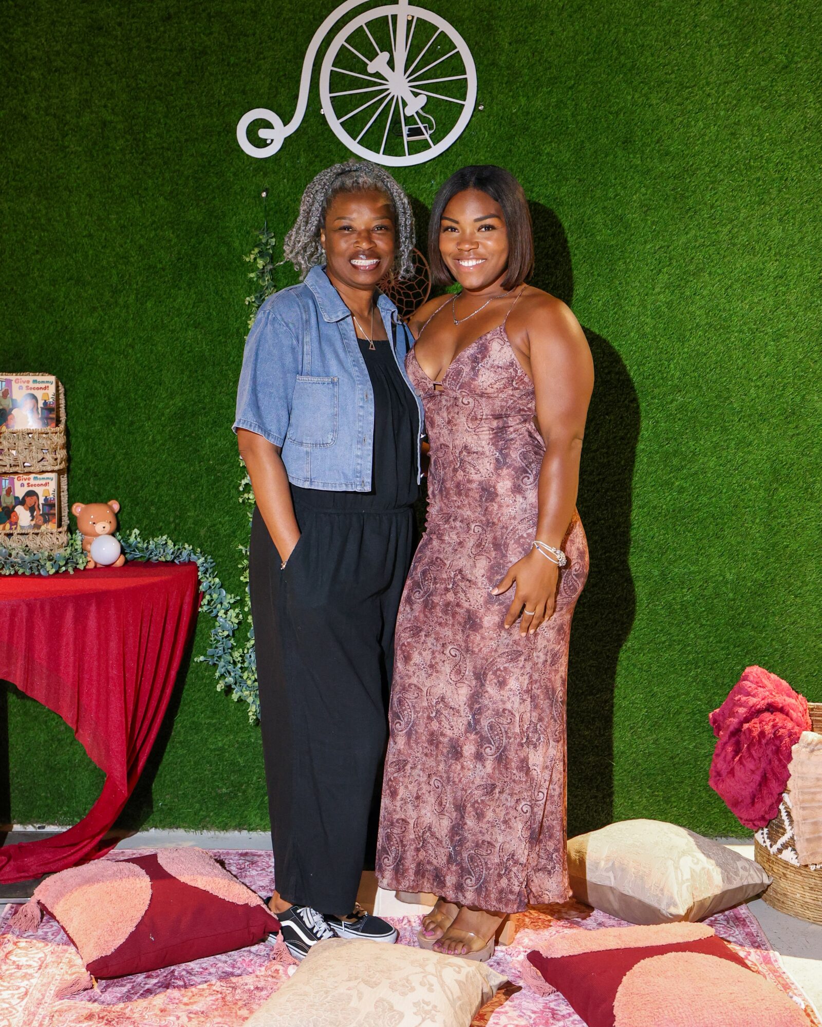 Two women stand together in front of a green wall with a bicycle decoration, smiling, with a table and pillows nearby.