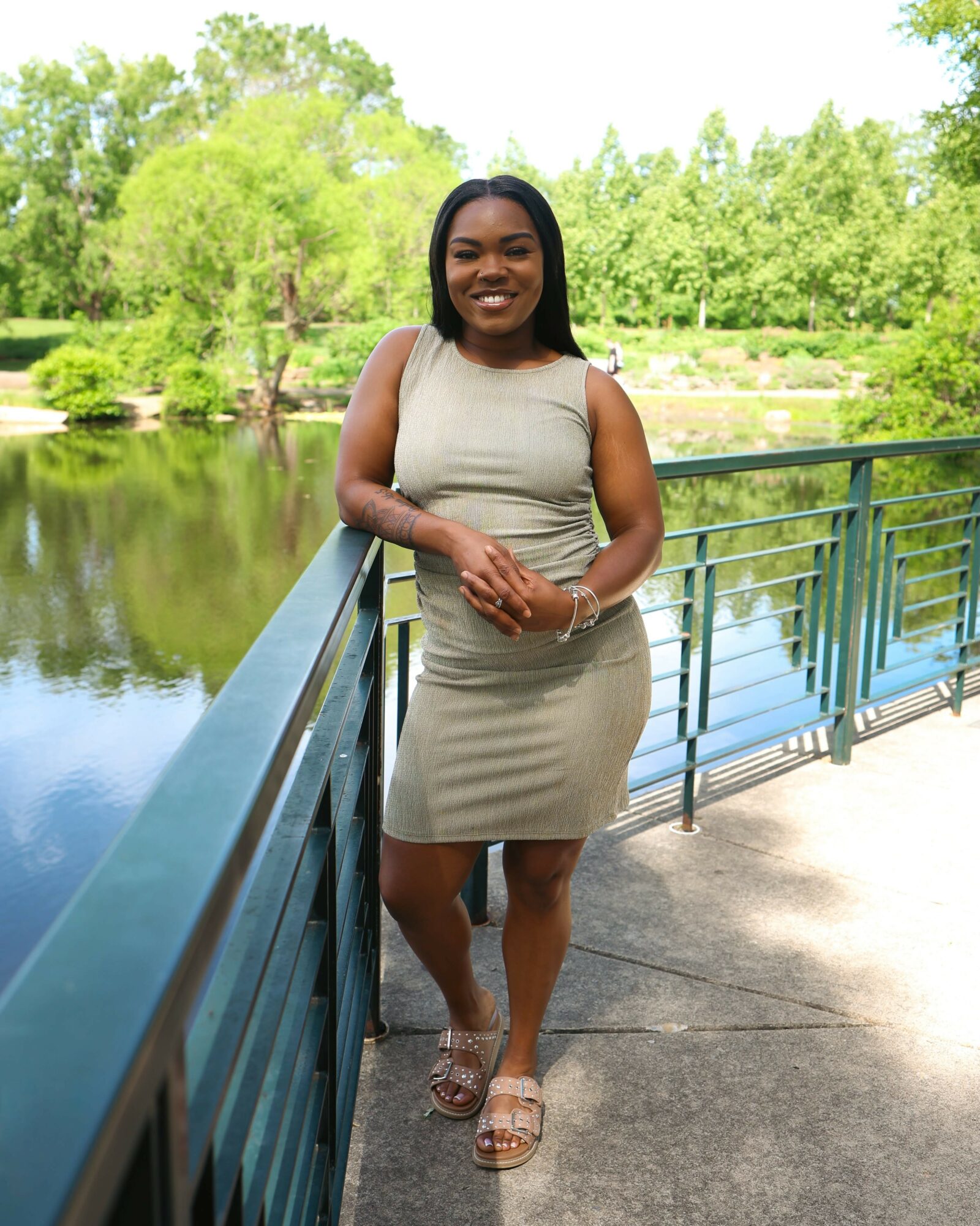 Woman standing on a bridge with a river and green trees in the background, smiling, wearing a beige dress and sandals.