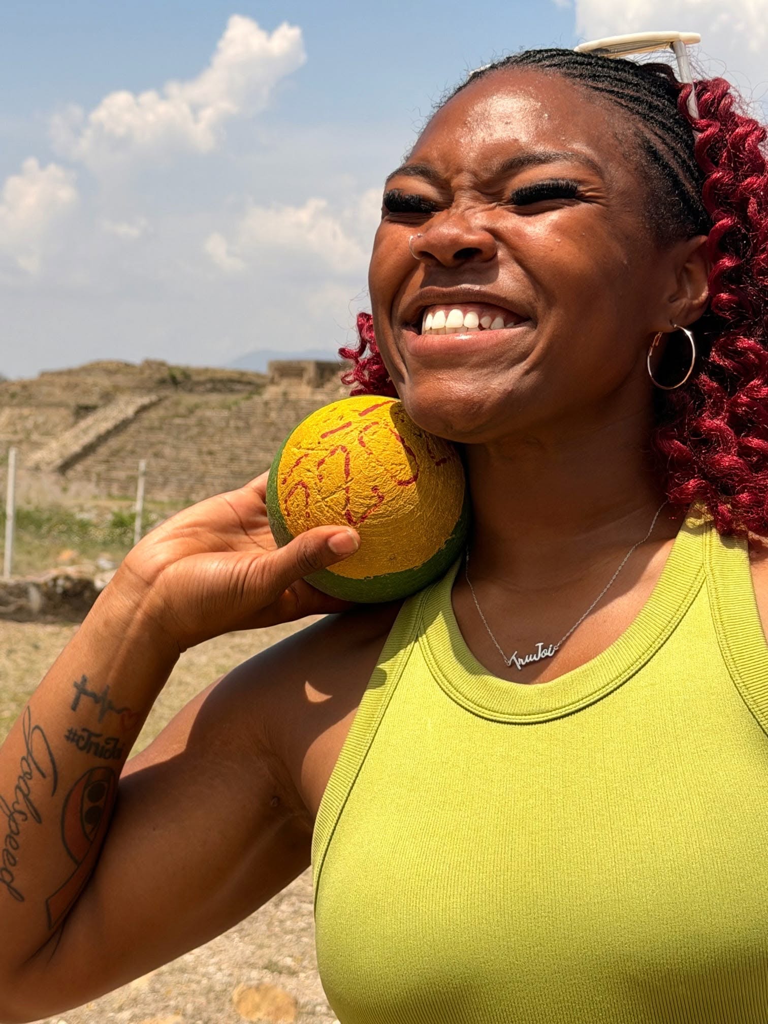 Woman smiling, holding a yellow ball with red patterns on her shoulder outdoors, with a cloudy sky and landscape in background.