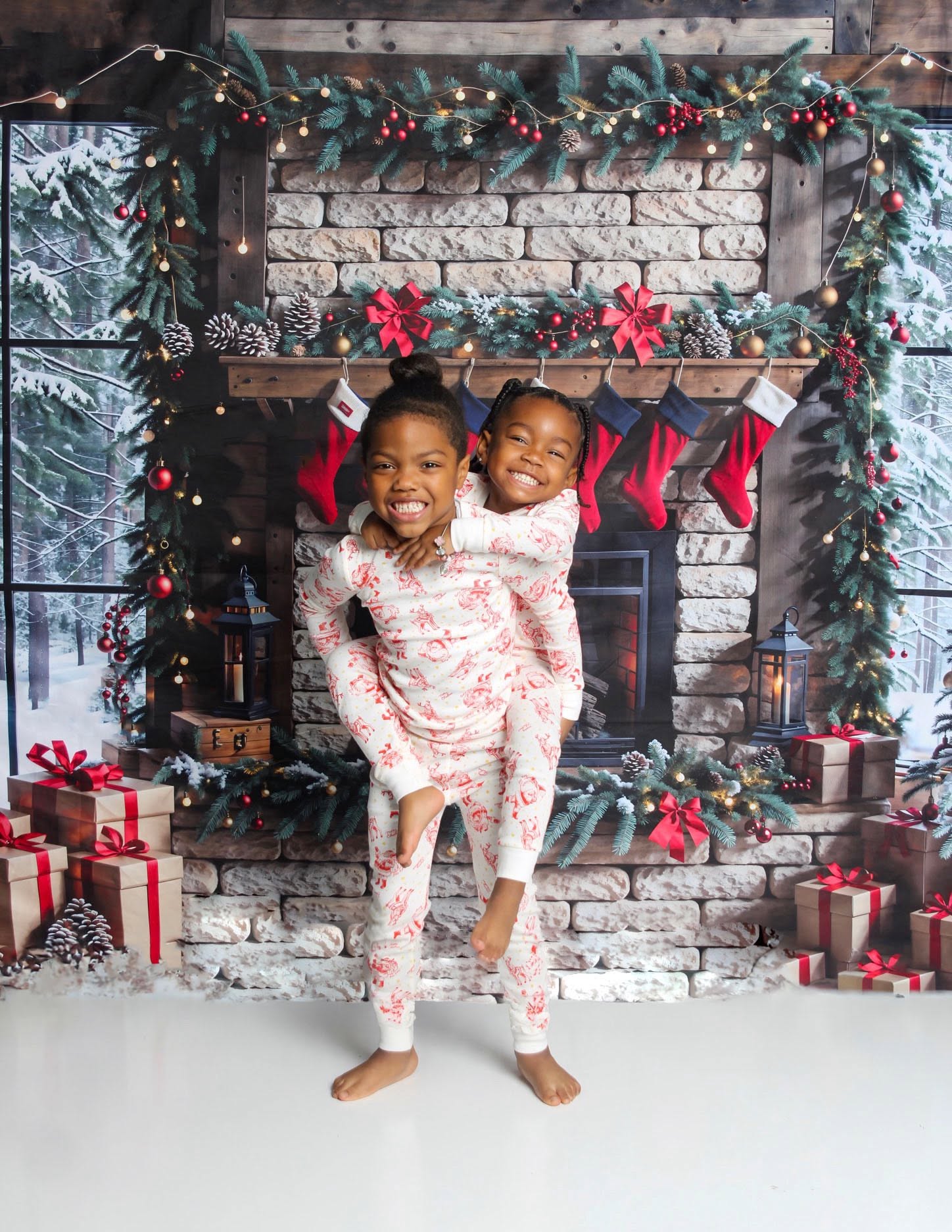 Two children in pajamas stand in front of a decorated fireplace with stockings and presents, smiling happily.