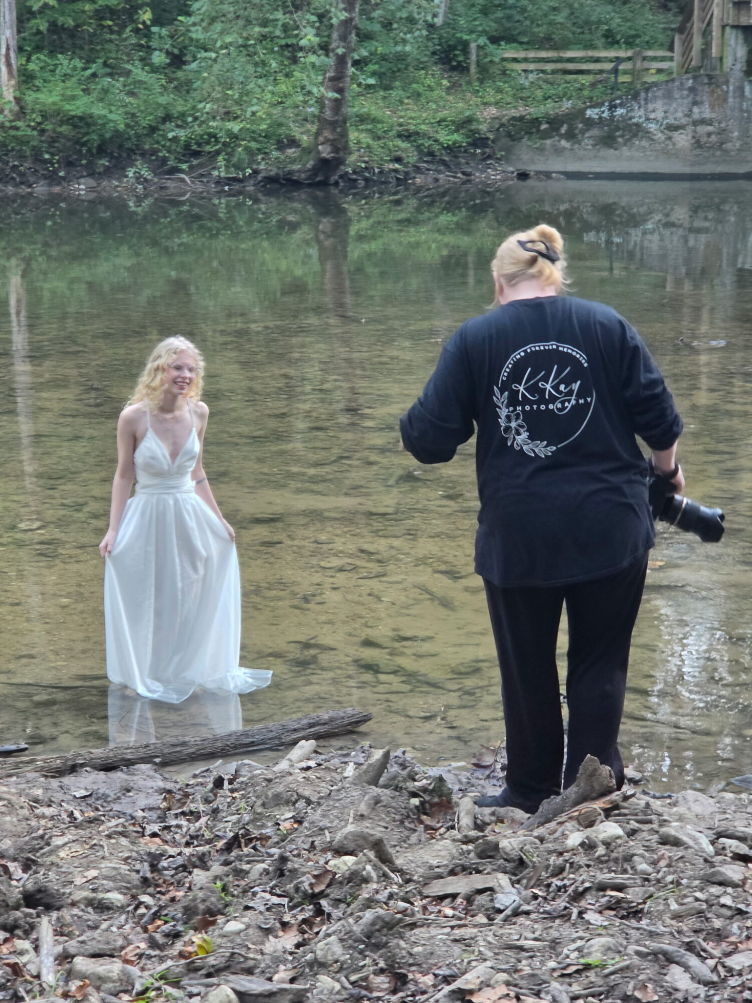 Person with blonde hair in a dark jacket holding a camera stands on rocky shore, facing a woman in a white dress in water.