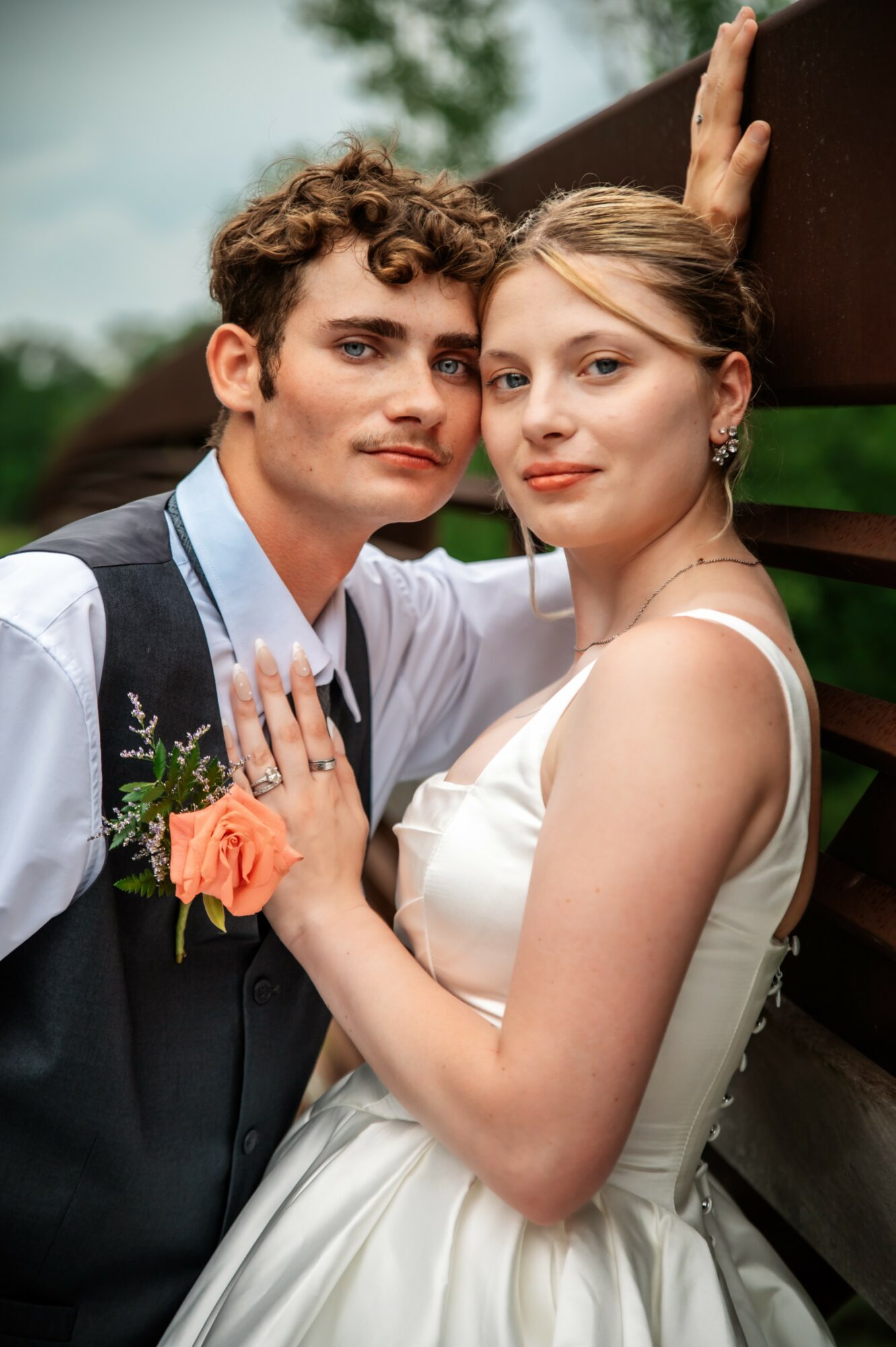 Young couple dressed in formal attire, close together outdoors, with a fence and trees in background.