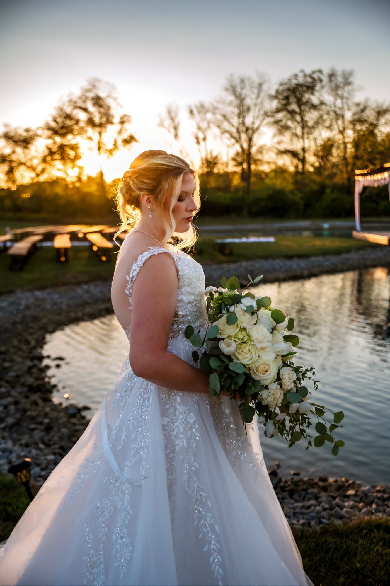 Bride in wedding dress holding a bouquet near a pond during sunset, trees in background.