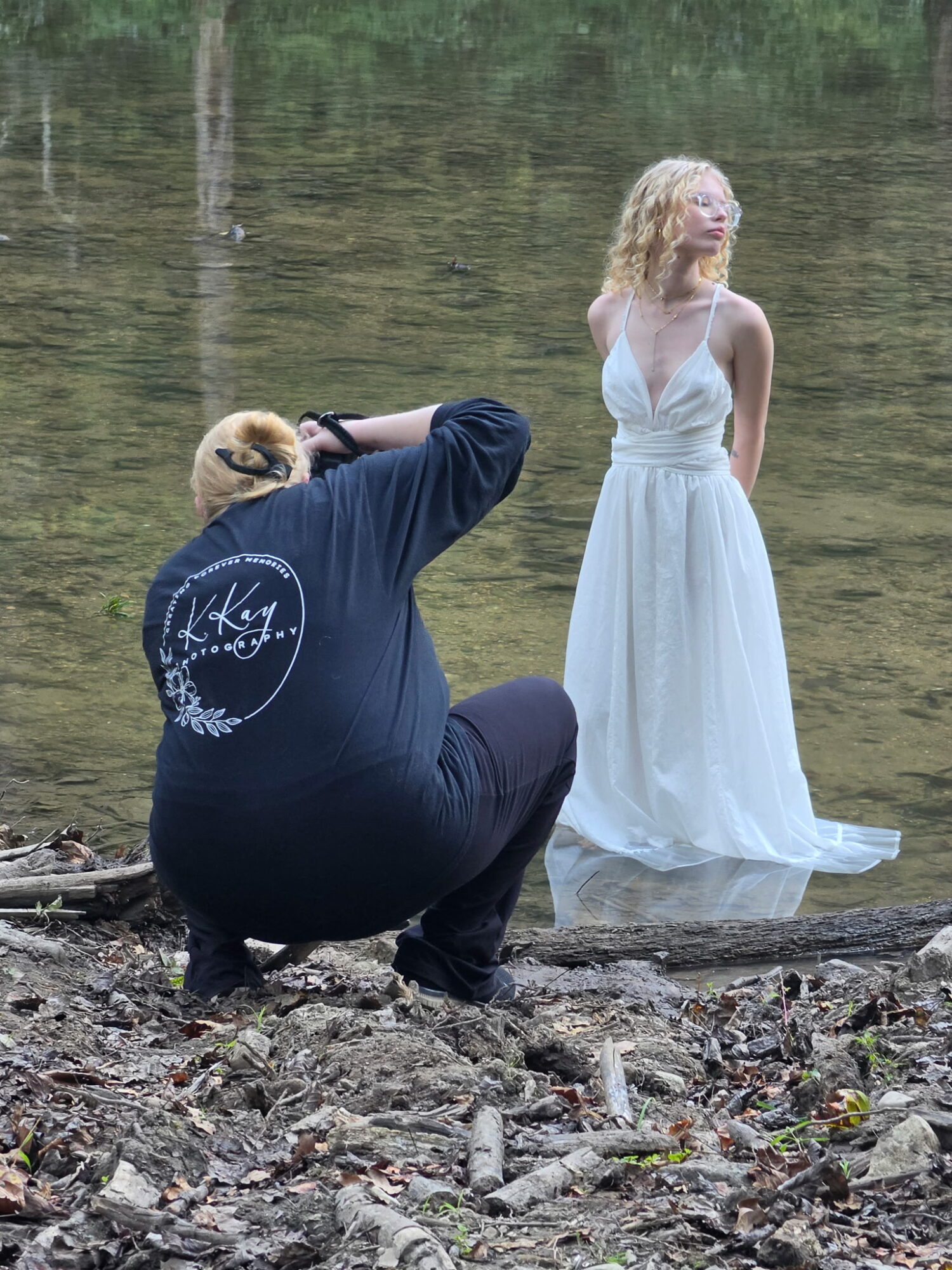 Photographer crouching on rocky shore taking picture of woman in white dress by water, trees in background.