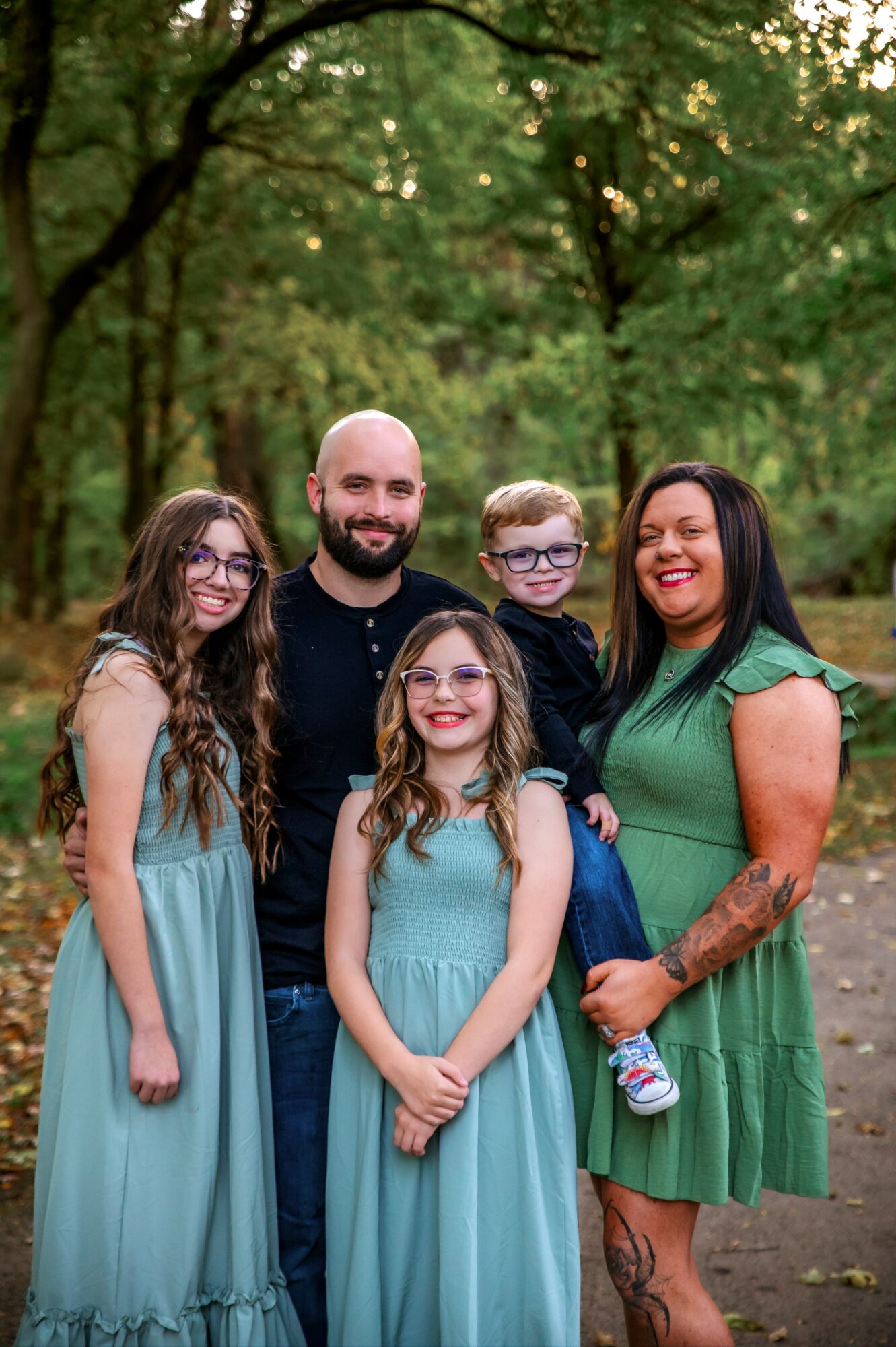 Family of five outdoors in a wooded area, smiling, with three children and two adults, all dressed in casual clothing.