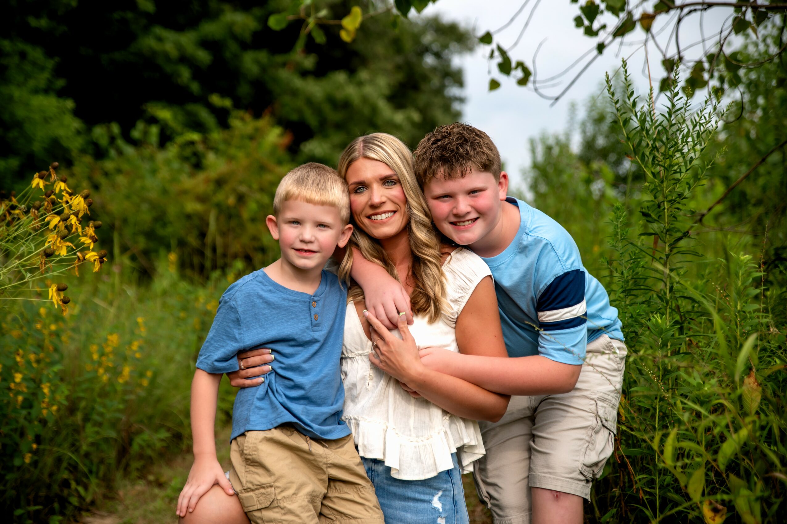 A woman with two boys outdoors surrounded by greenery, smiling and hugging each other.
