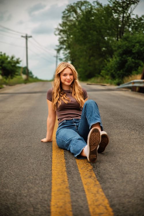 Young woman sitting on a road with trees and power lines in the background, smiling, wearing a brown shirt and jeans.