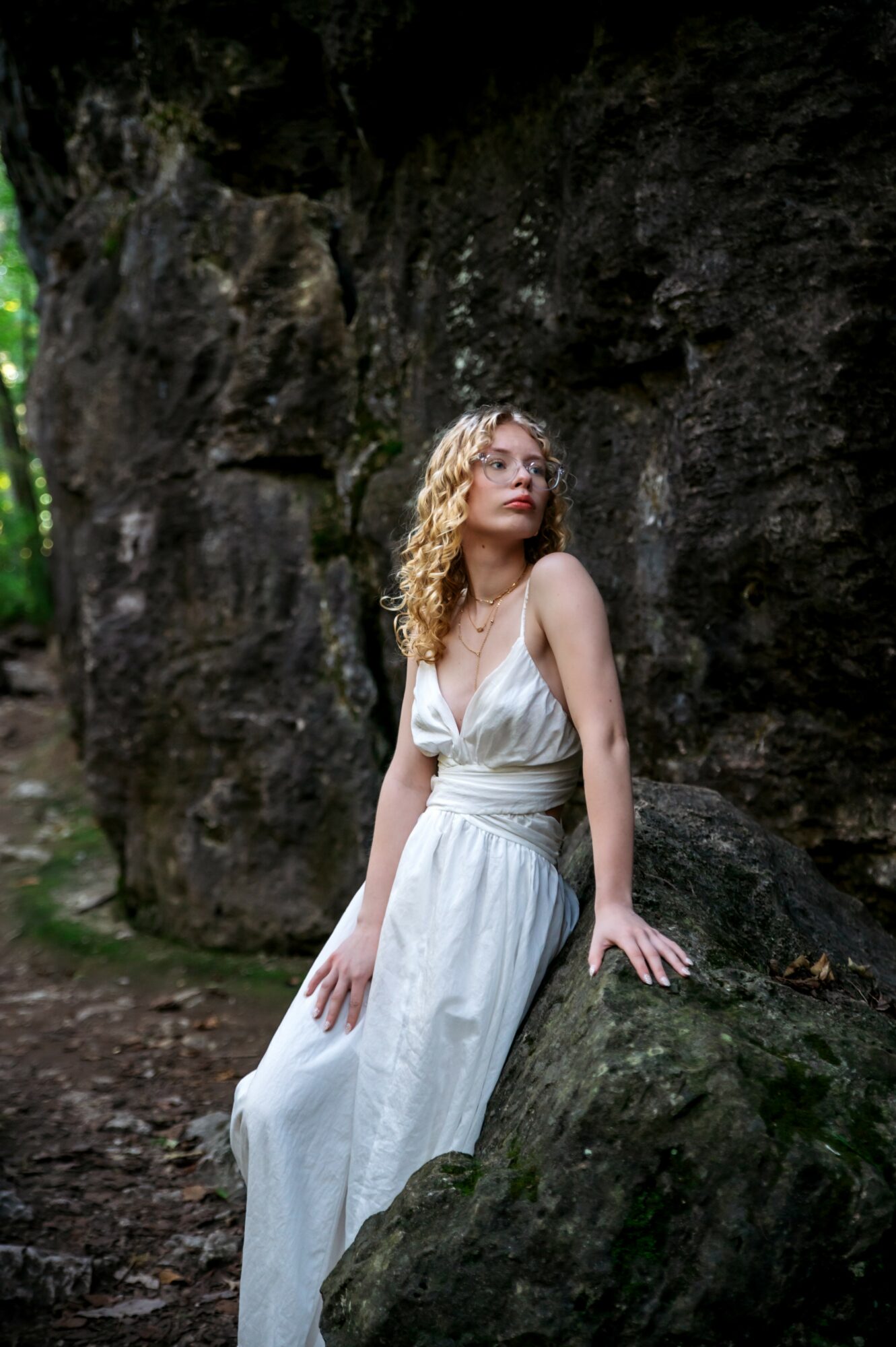 Woman with curly hair in a white dress sitting on a large rock in a forest setting.
