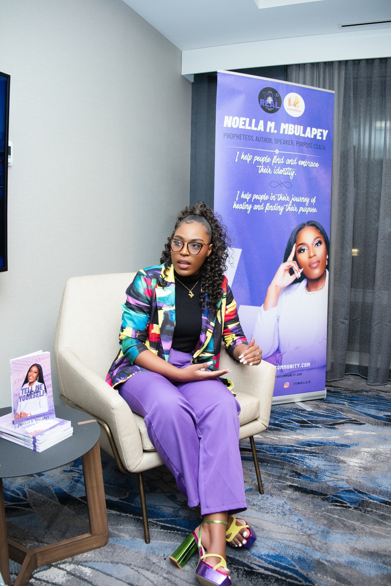Woman with curly hair and glasses sitting on a beige chair, wearing a colorful blazer and purple pants, in a room with a banner and a table.