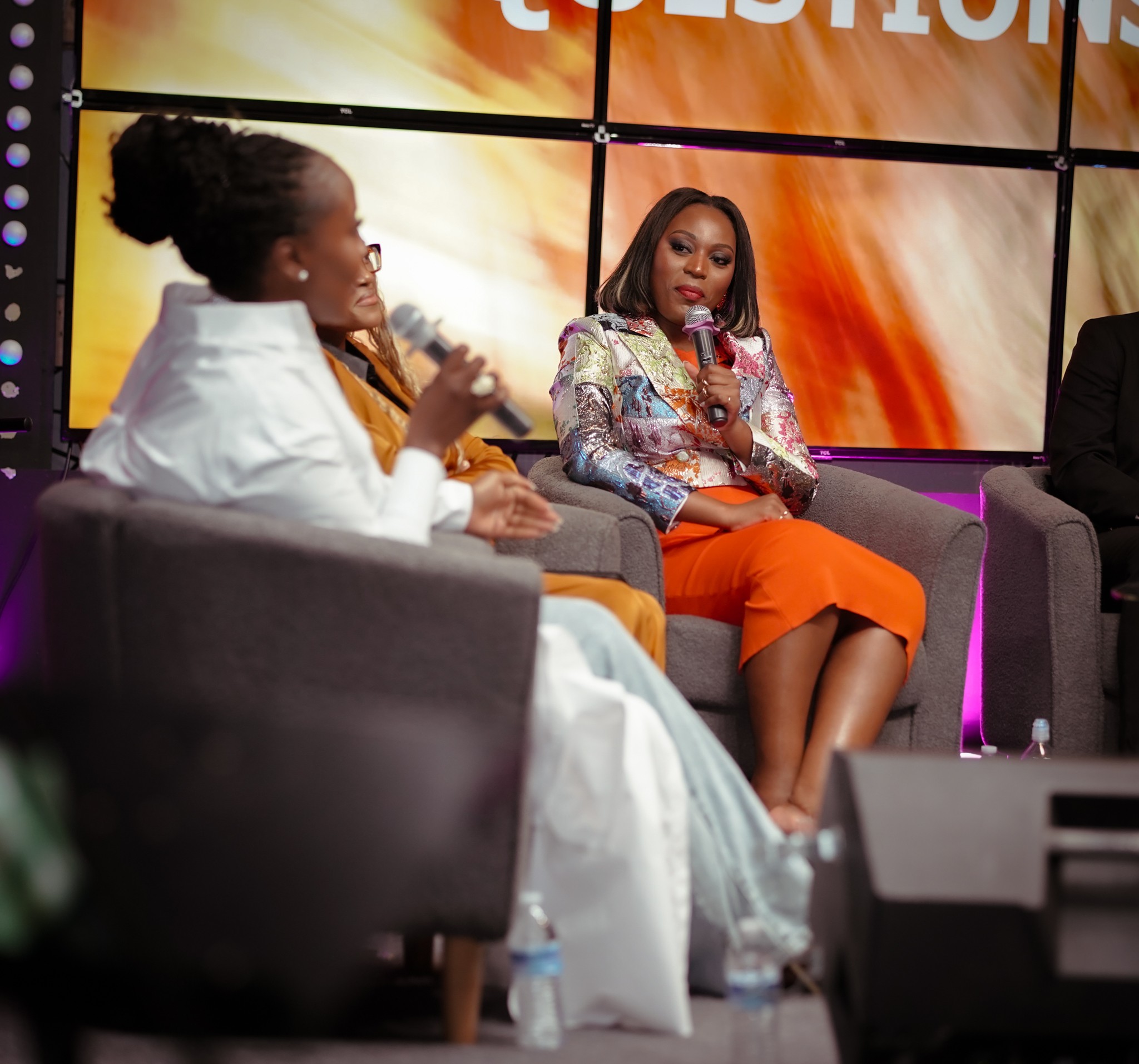 Two women seated on stage, one speaking into a microphone, the other listening, with a large screen behind them.