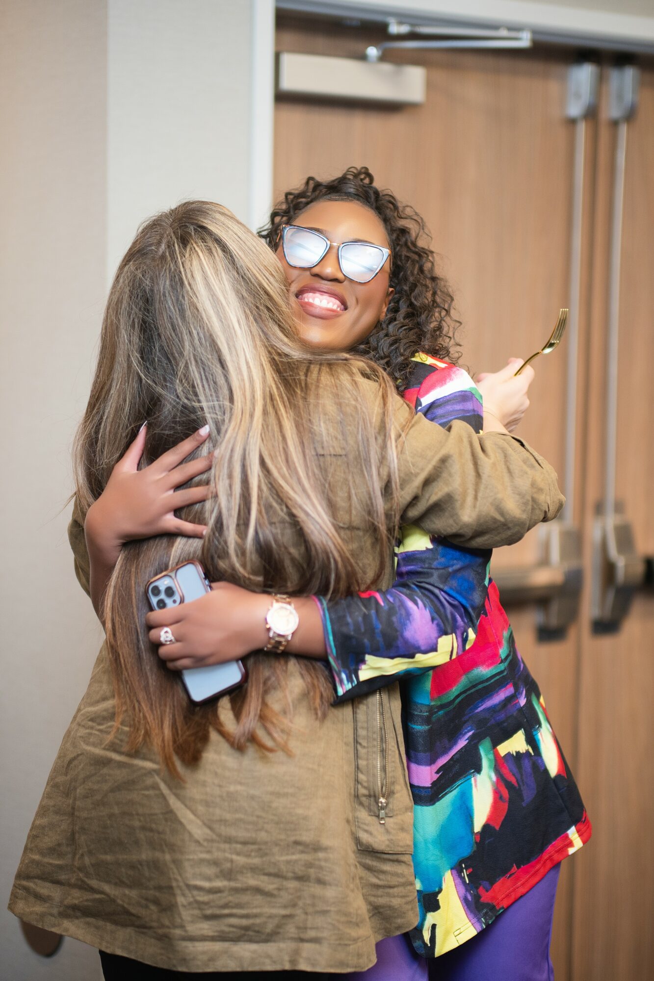 Two women hugging, one with curly hair and sunglasses, the other with long straight hair, in a room with wooden doors