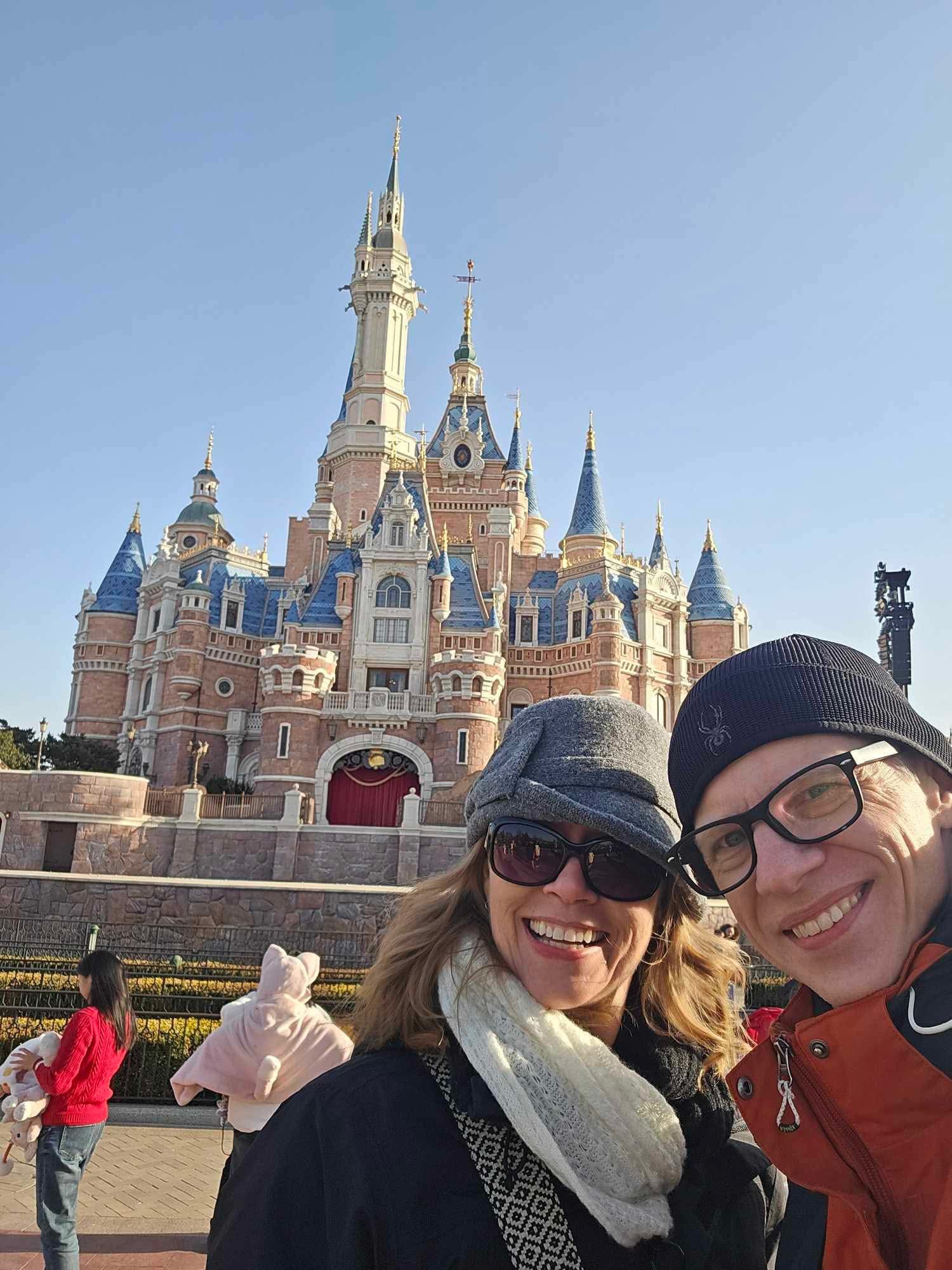 Two smiling women wearing hats and glasses in front of a castle with tall spires and turrets, clear blue sky.