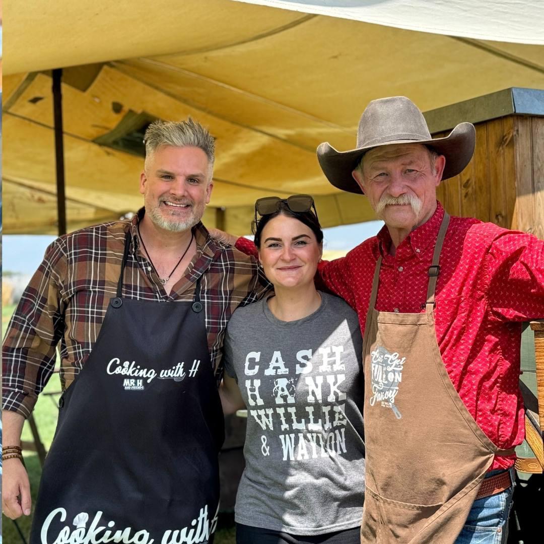 Three people smiling outdoors under a yellow canopy, two men and a woman, one man wearing a cowboy hat, all in casual clothing.