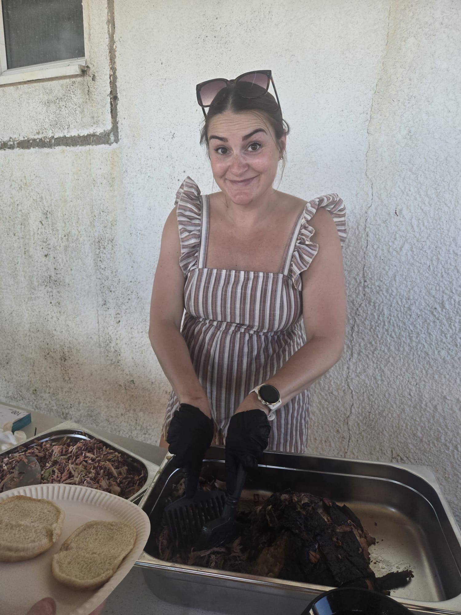 Young woman with cat ears headband, smiling, wearing striped dress, standing behind a table with food, against a white wall.