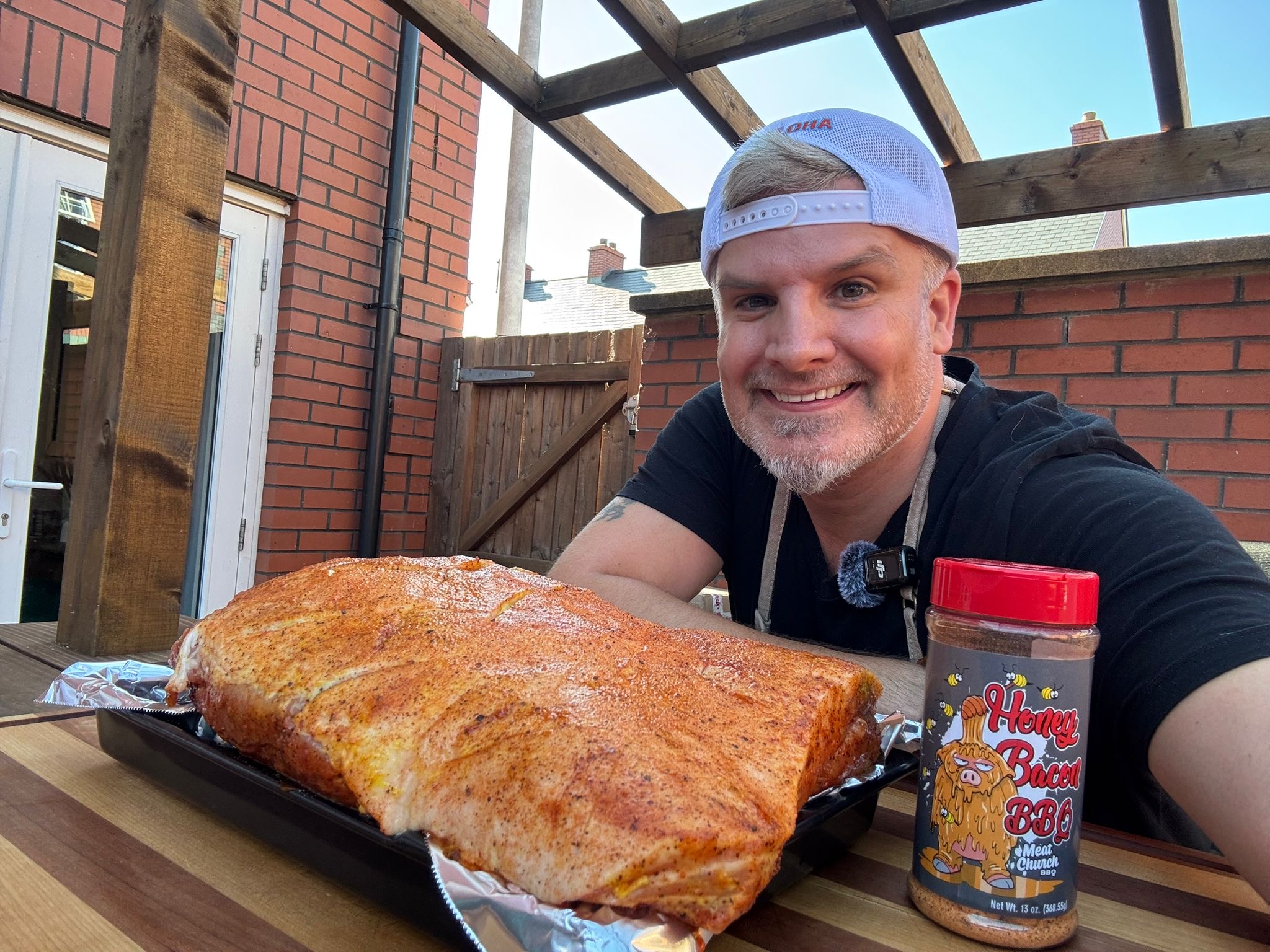 Smiling man with a cap and black shirt sits at a table with a large piece of cooked salmon, outdoor setting with brick wall and wooden structure