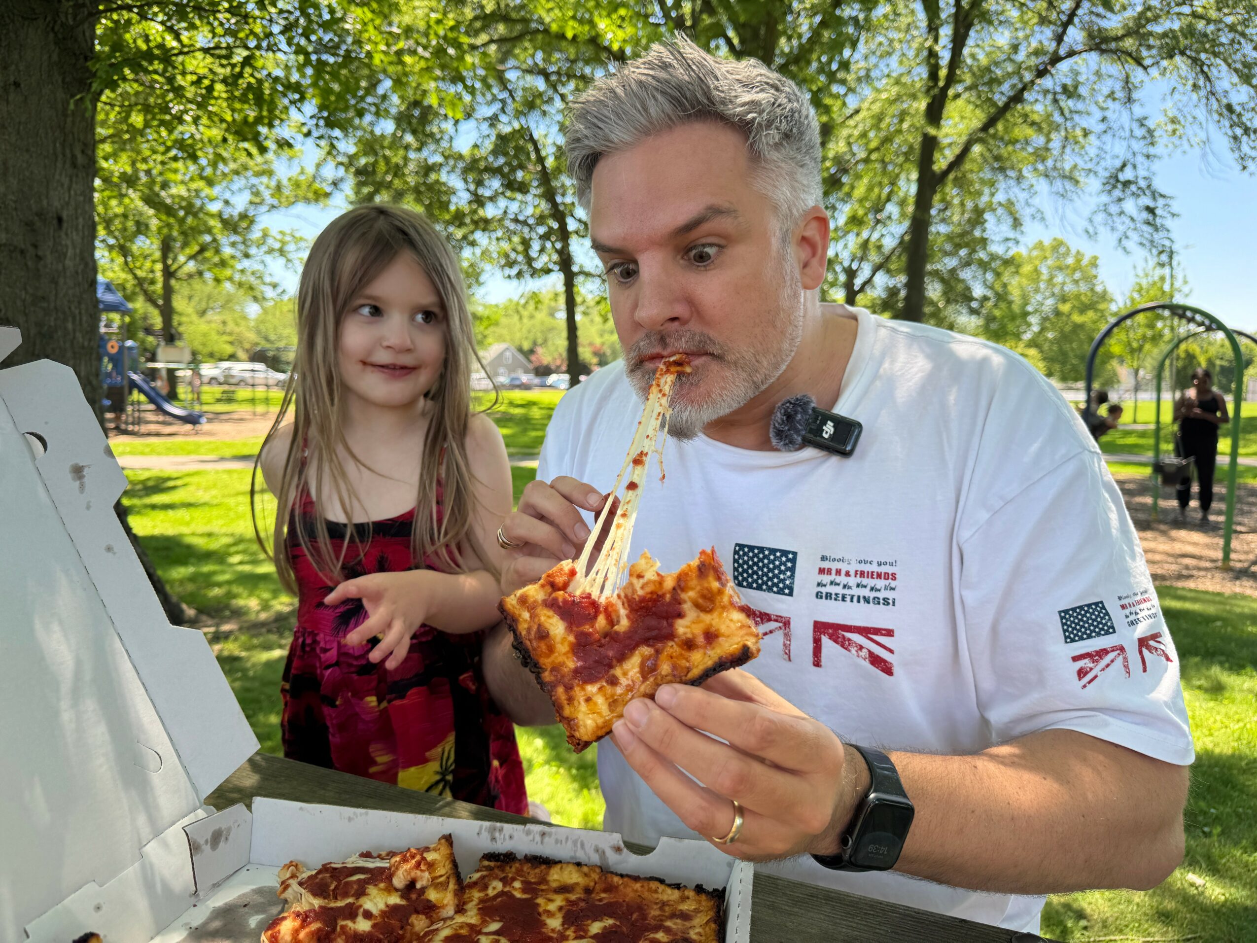 Man and young girl outdoors, man eating pizza, girl watching, trees and park in background.