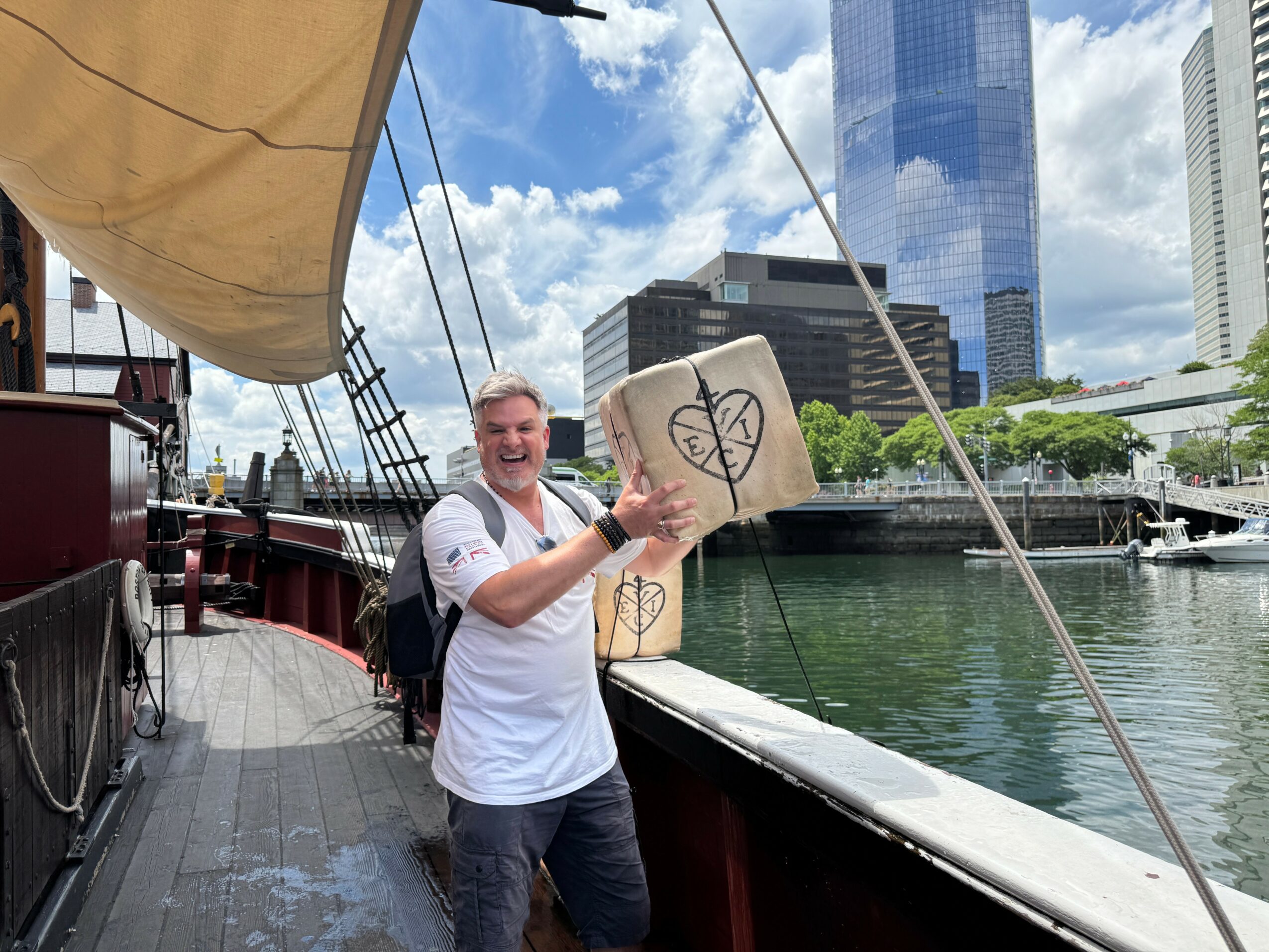 Man smiling on a boat with a city skyline and water in the background, holding a cardboard box with a drawing. Word count: 24