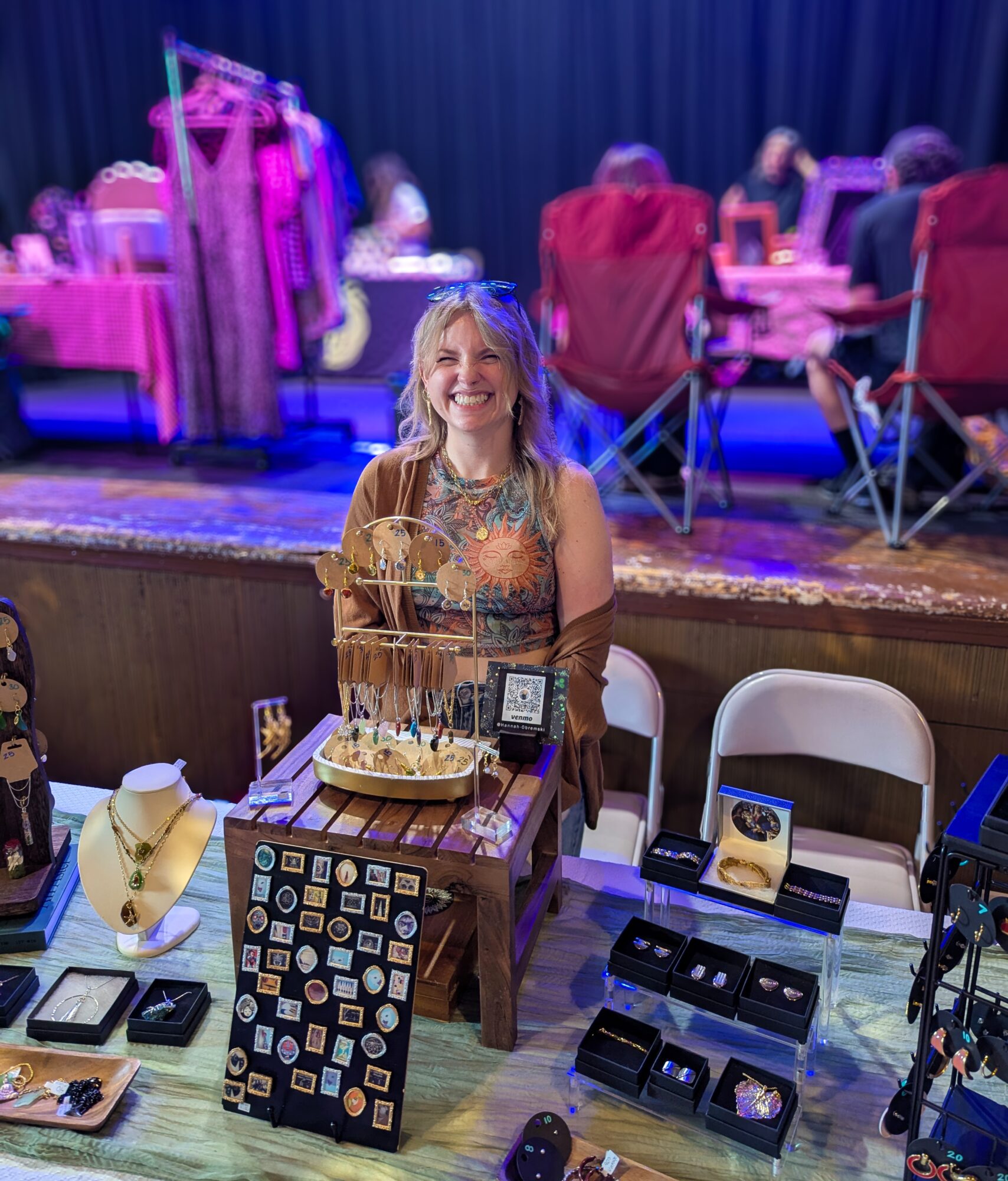 Person seated behind a table displaying jewelry and small items at an indoor event.