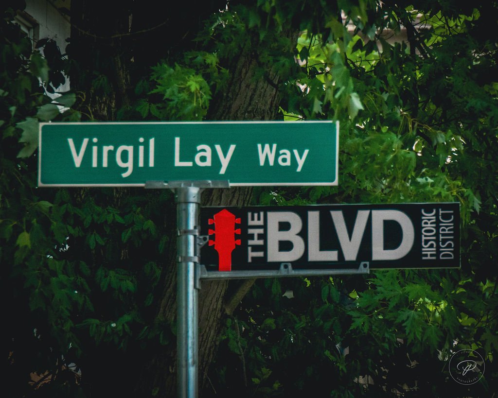 Street signs for Virgil Lay Way and The BLVD Historic District amid leafy trees.