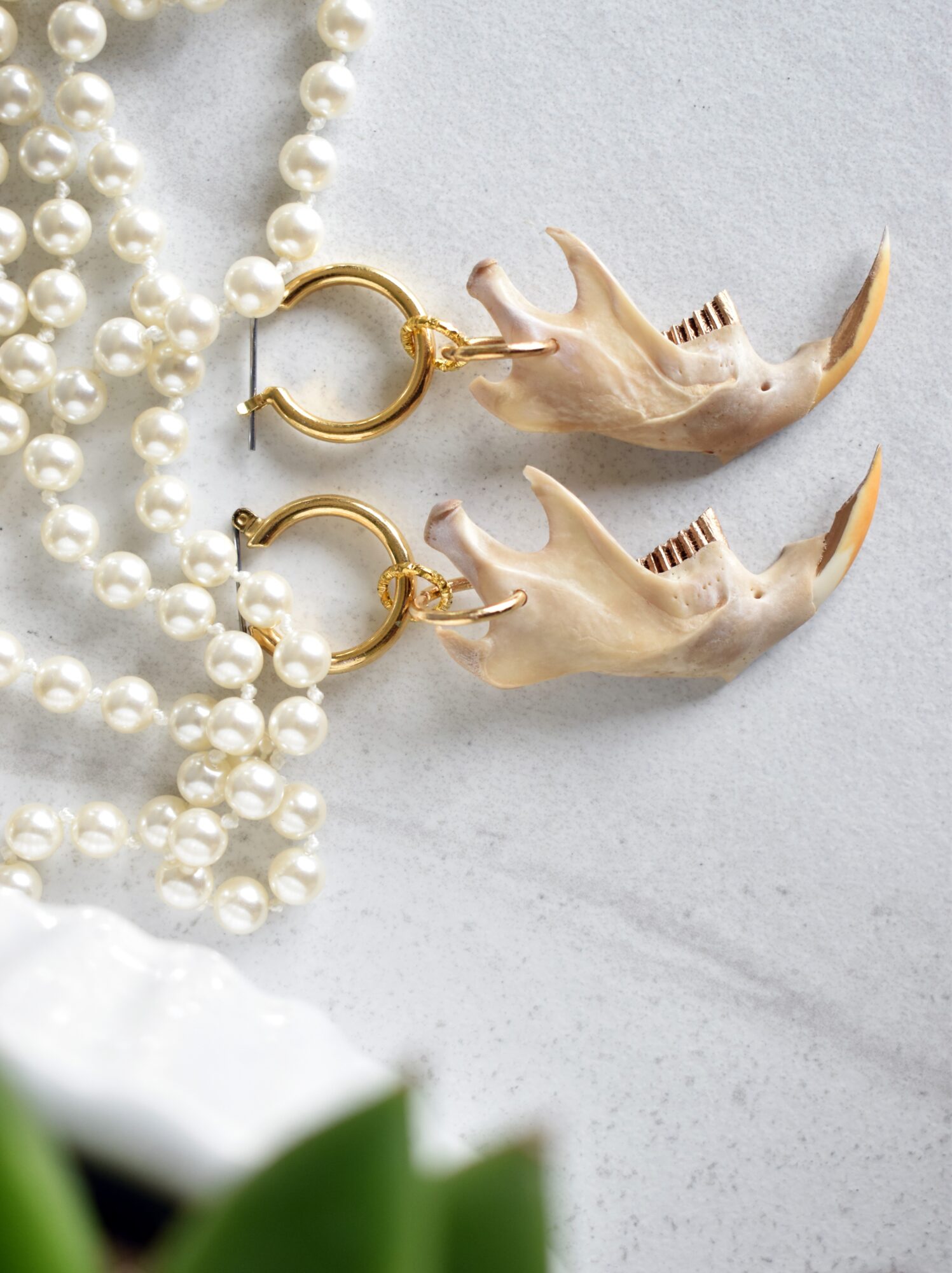 Two gold hoop earrings with bird-shaped pendants lie beside strands of white pearls and green leaves.