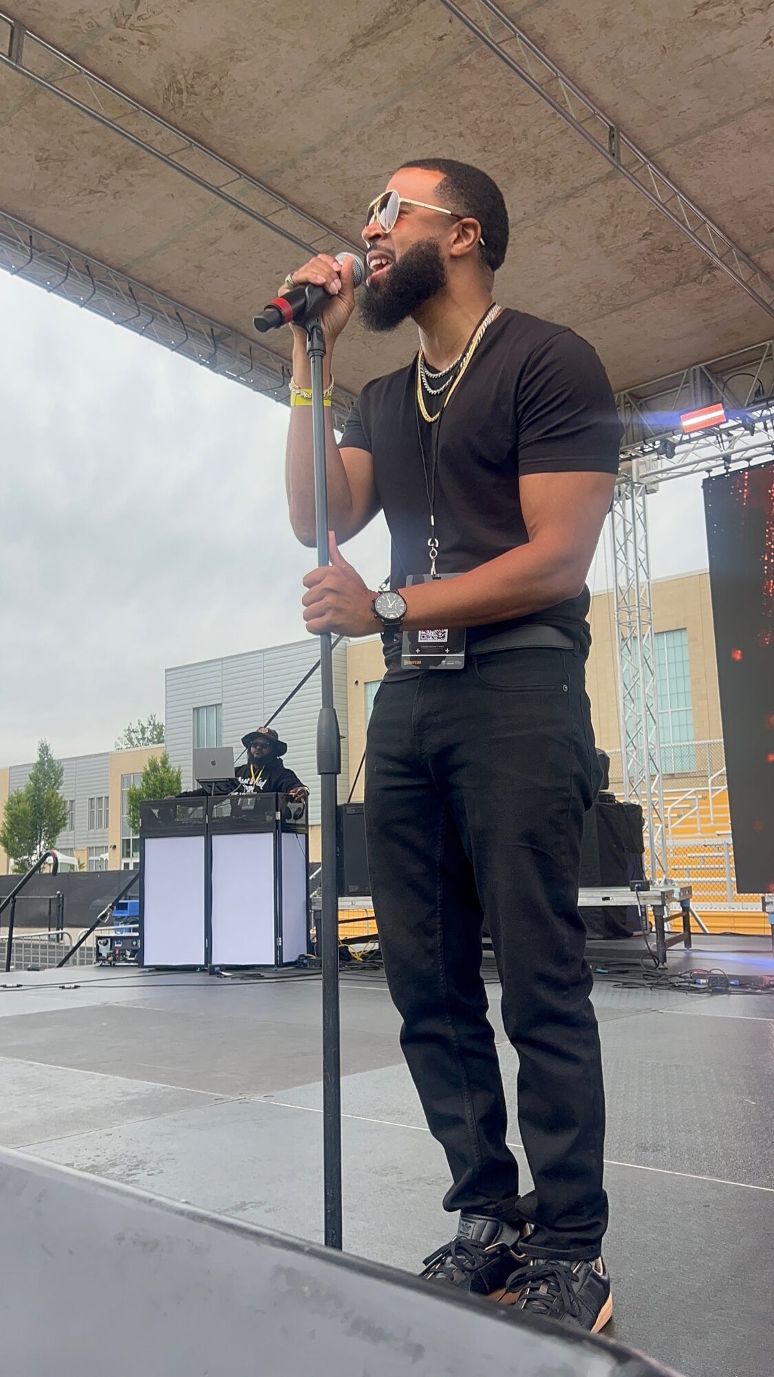 A person wearing sunglasses and black clothing sings into a microphone on an outdoor stage.