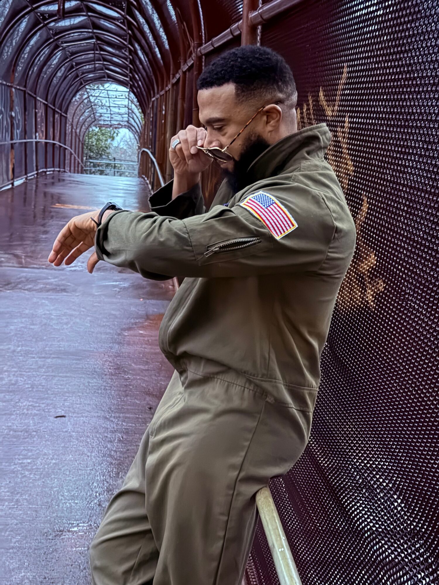 Person in an olive flight suit leaning against a mesh fence inside a curved metal walkway.