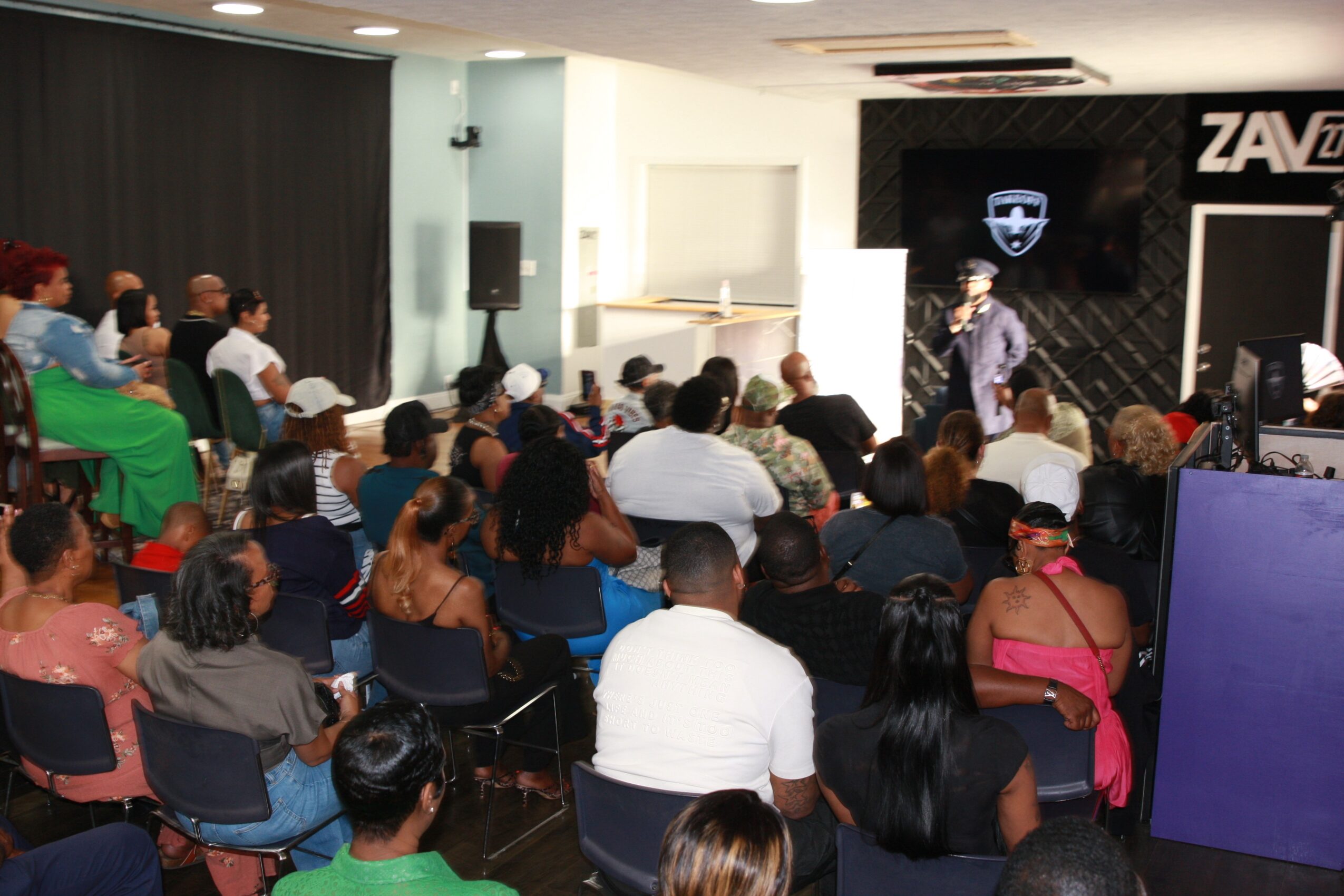 Audience seated in a room facing a presenter and a screen with logos.