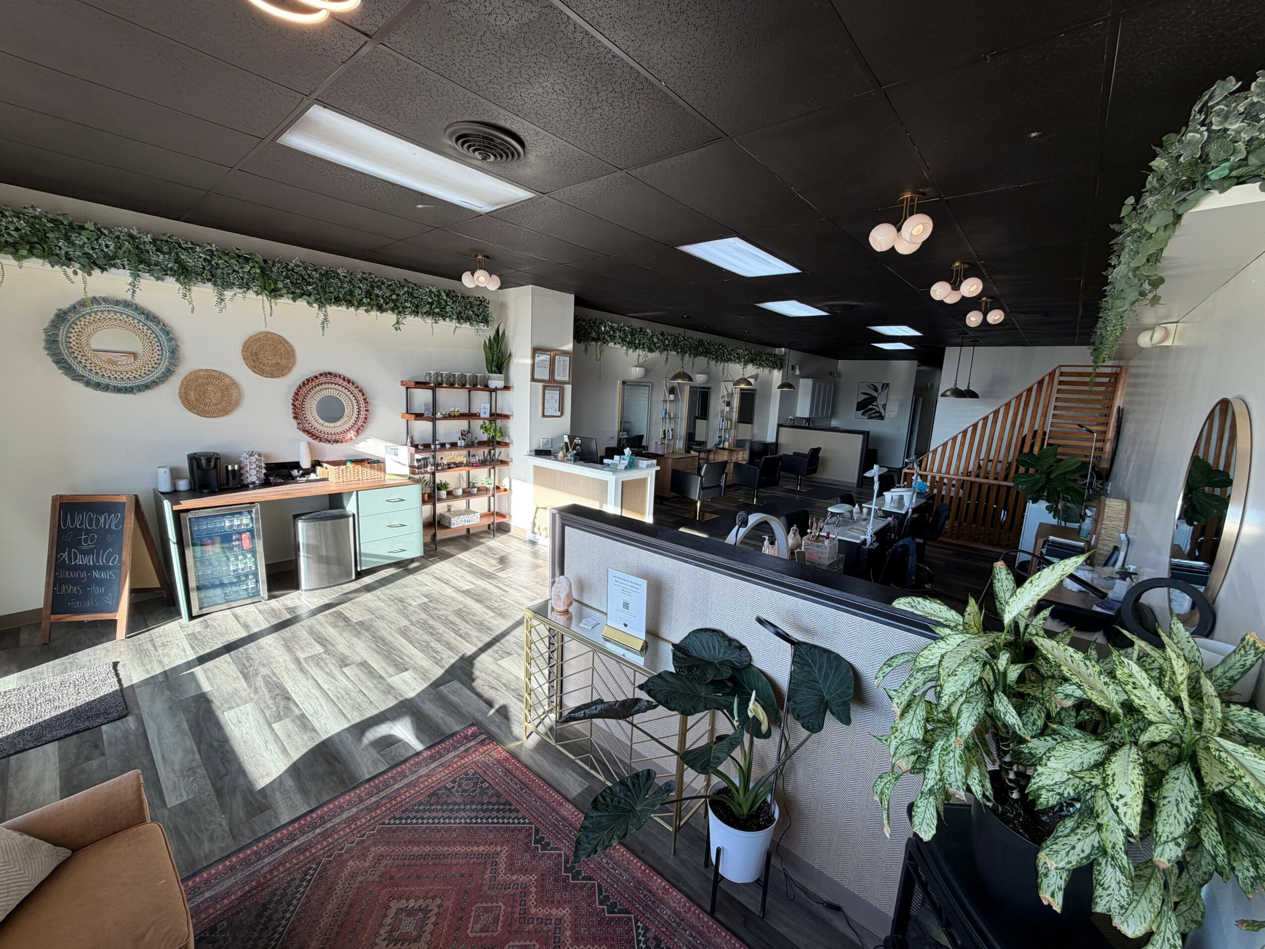 Interior of a cafe with plants, wall decorations, a counter, and seating area, illuminated by ceiling lights and natural light.