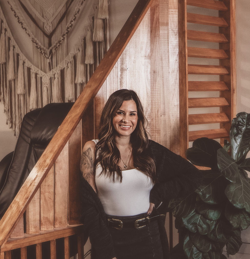 Smiling woman with long dark hair, wearing a white top and black jacket, sitting indoors near wooden decor and plants.