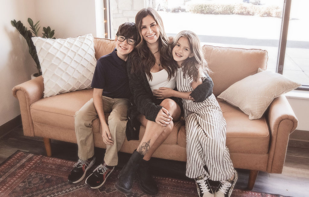 Three people sitting on a sofa, smiling, in a bright room with large windows and pillows.