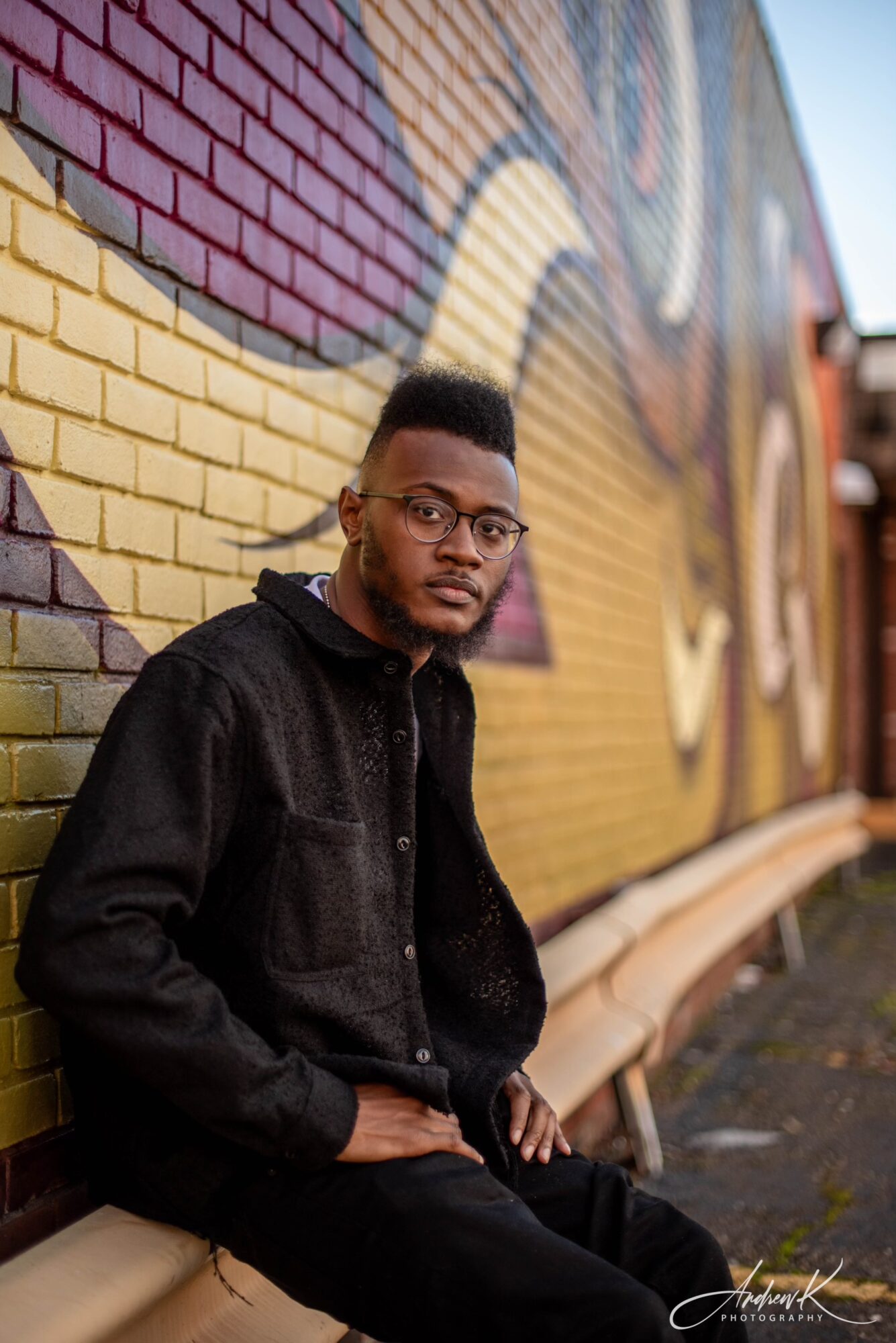 Young man with glasses and a black jacket leaning against a colorful graffiti wall.
