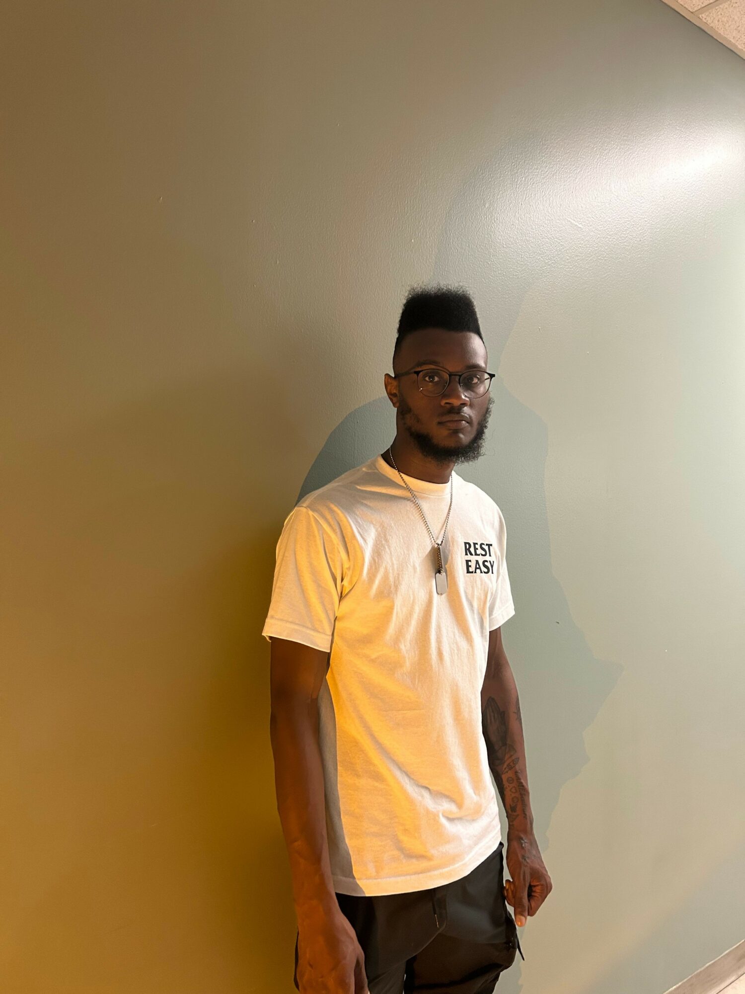 A man with glasses and a beard wearing a white T-shirt and dark pants stands against a plain wall.
