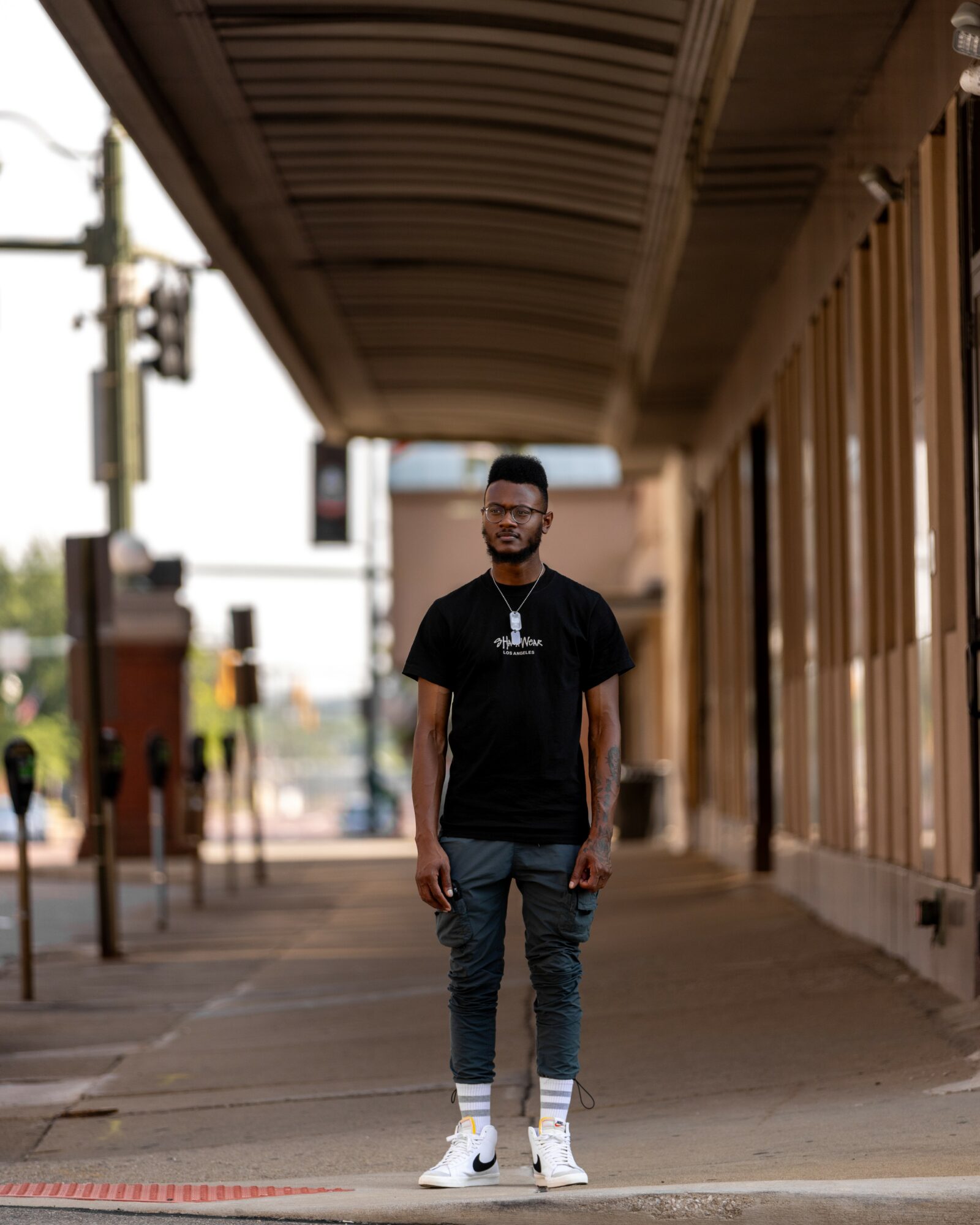 Young man standing on sidewalk under a covered walkway, wearing a black t-shirt, jeans, and white sneakers, smiling.