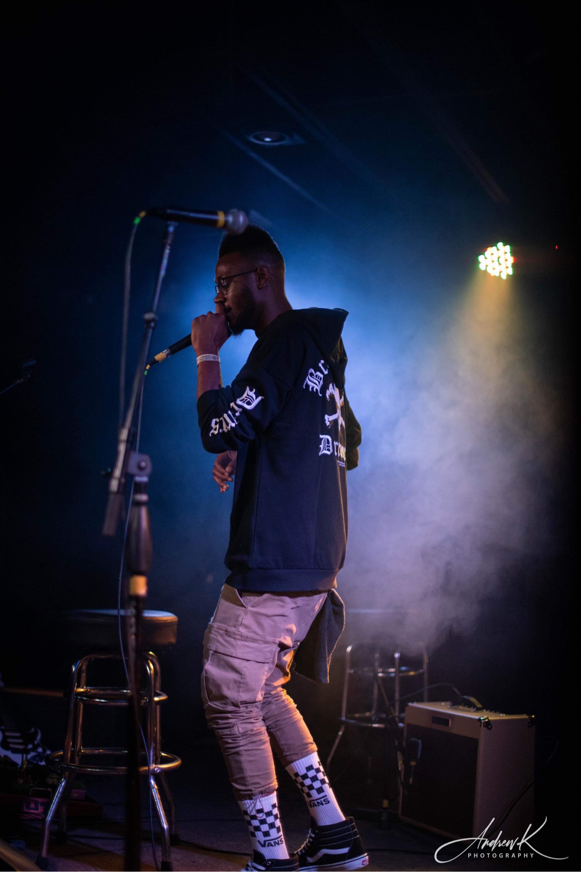 Person singing into microphone on stage with fog and bright light behind, wearing hoodie, pants, and checkered socks.
