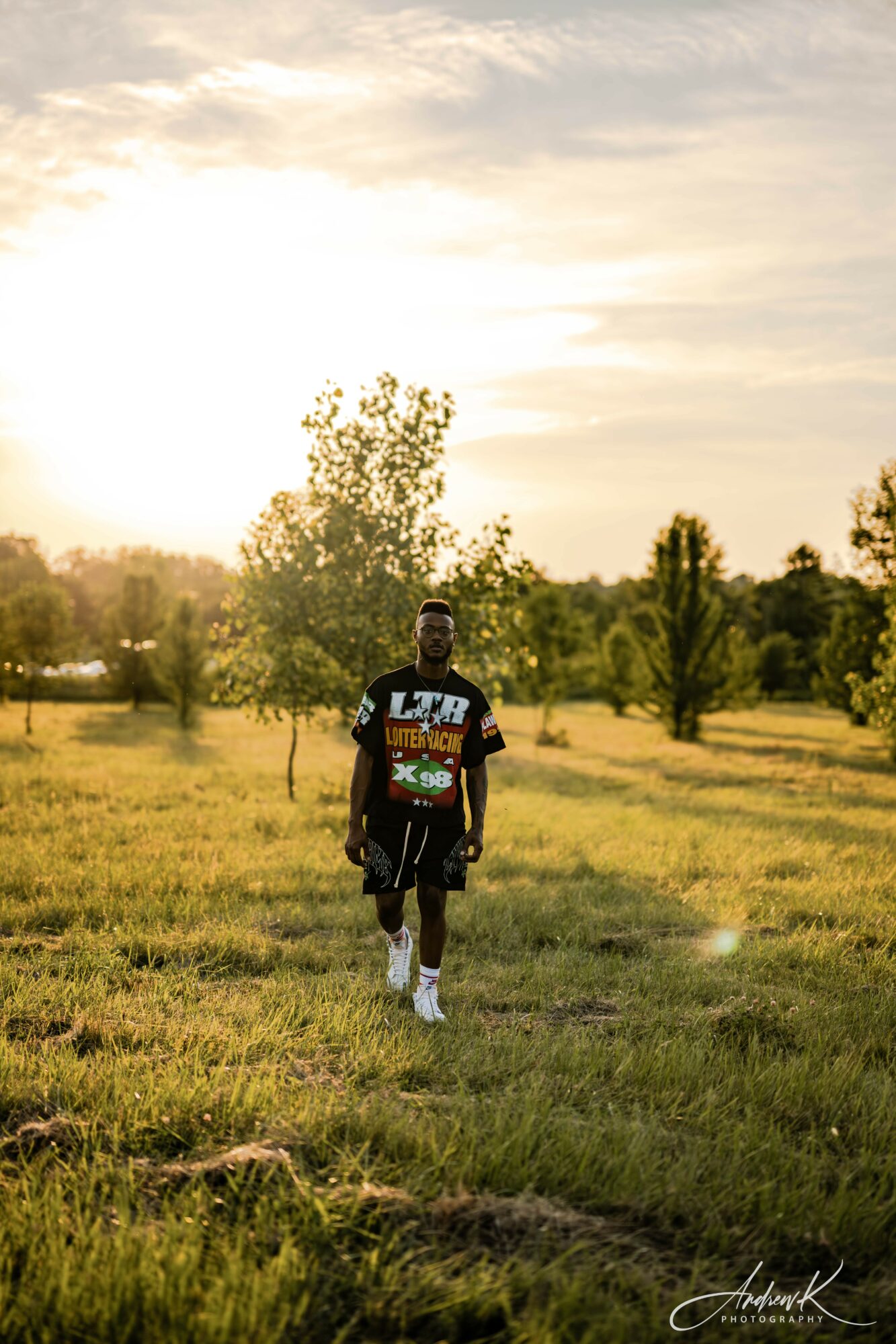 Person walking in a grassy field with trees and a sunset sky in the background.