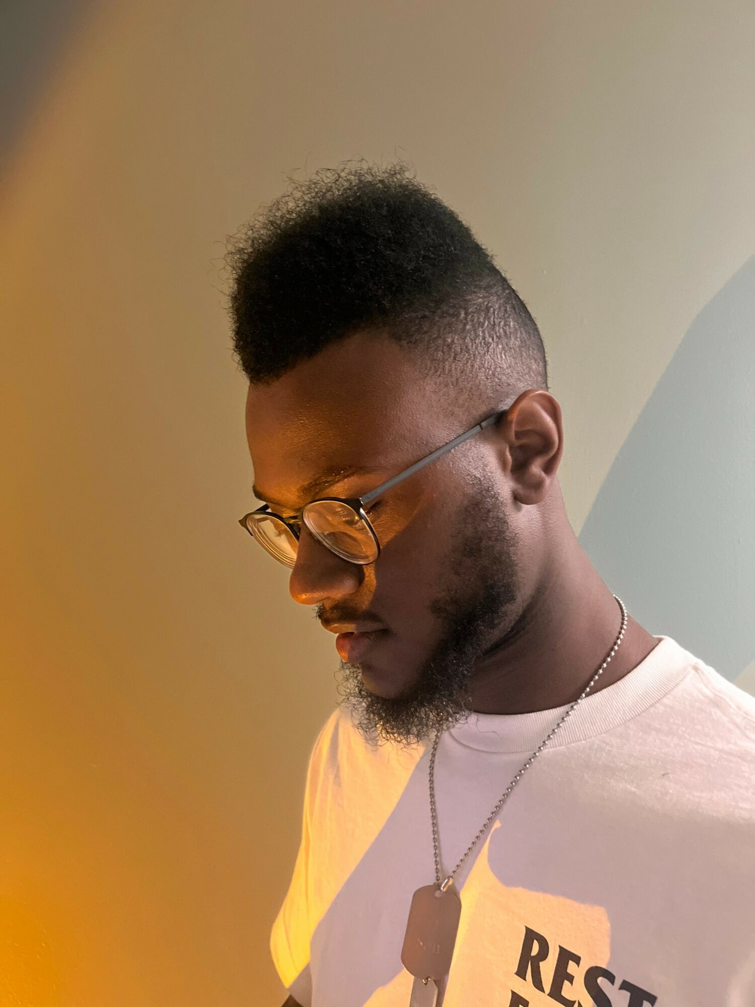Young man with glasses and a beard, wearing a white shirt, looking down, with a neutral background.