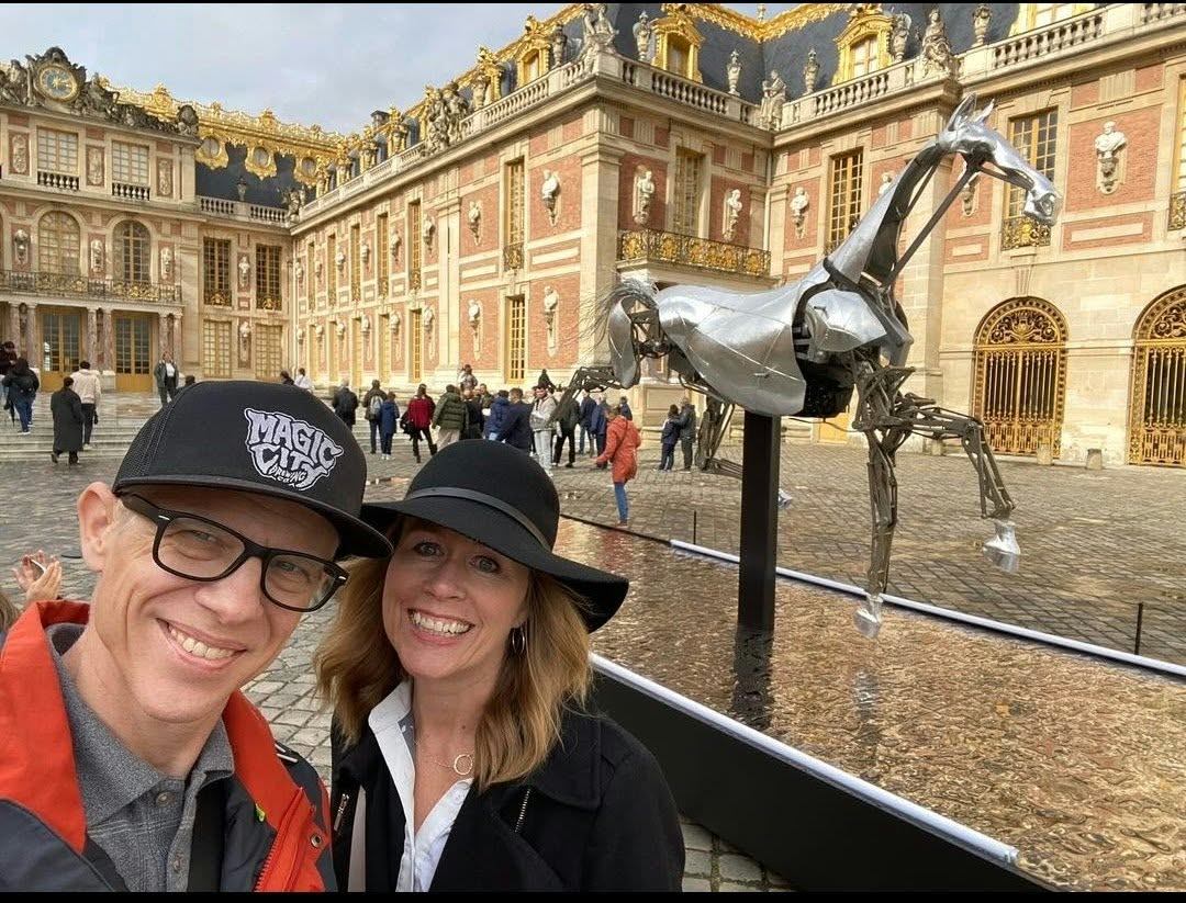 Two people smiling in front of a horse sculpture outside a grand historic building, with other visitors in the background.