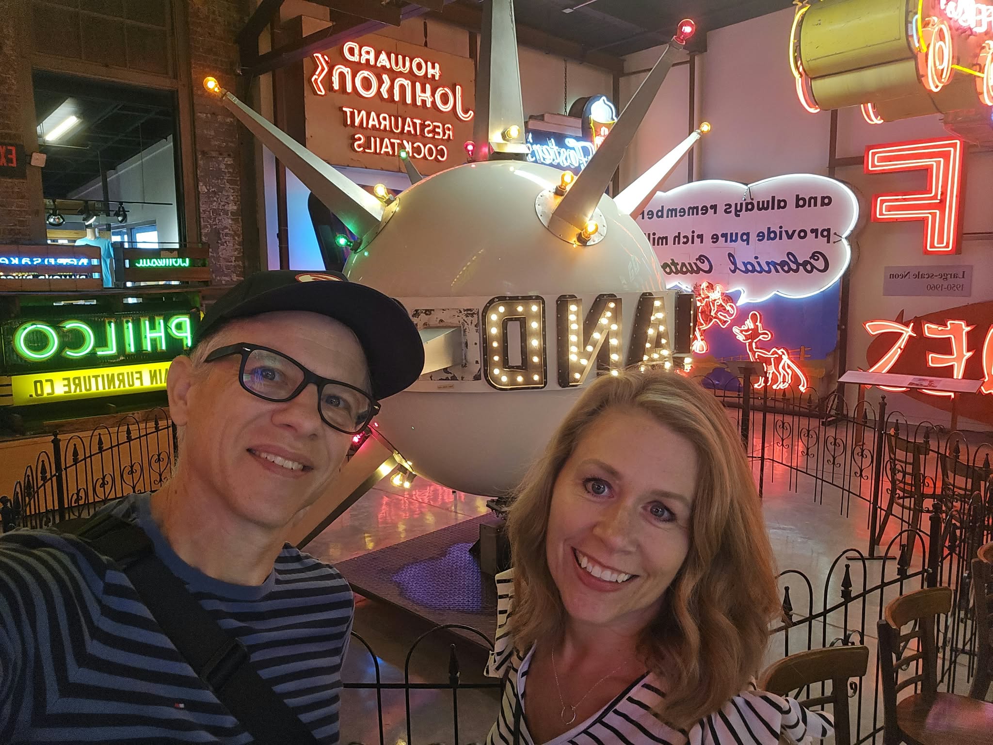 Two smiling people pose in front of a large unicorn sculpture with colorful neon signs in the background.
