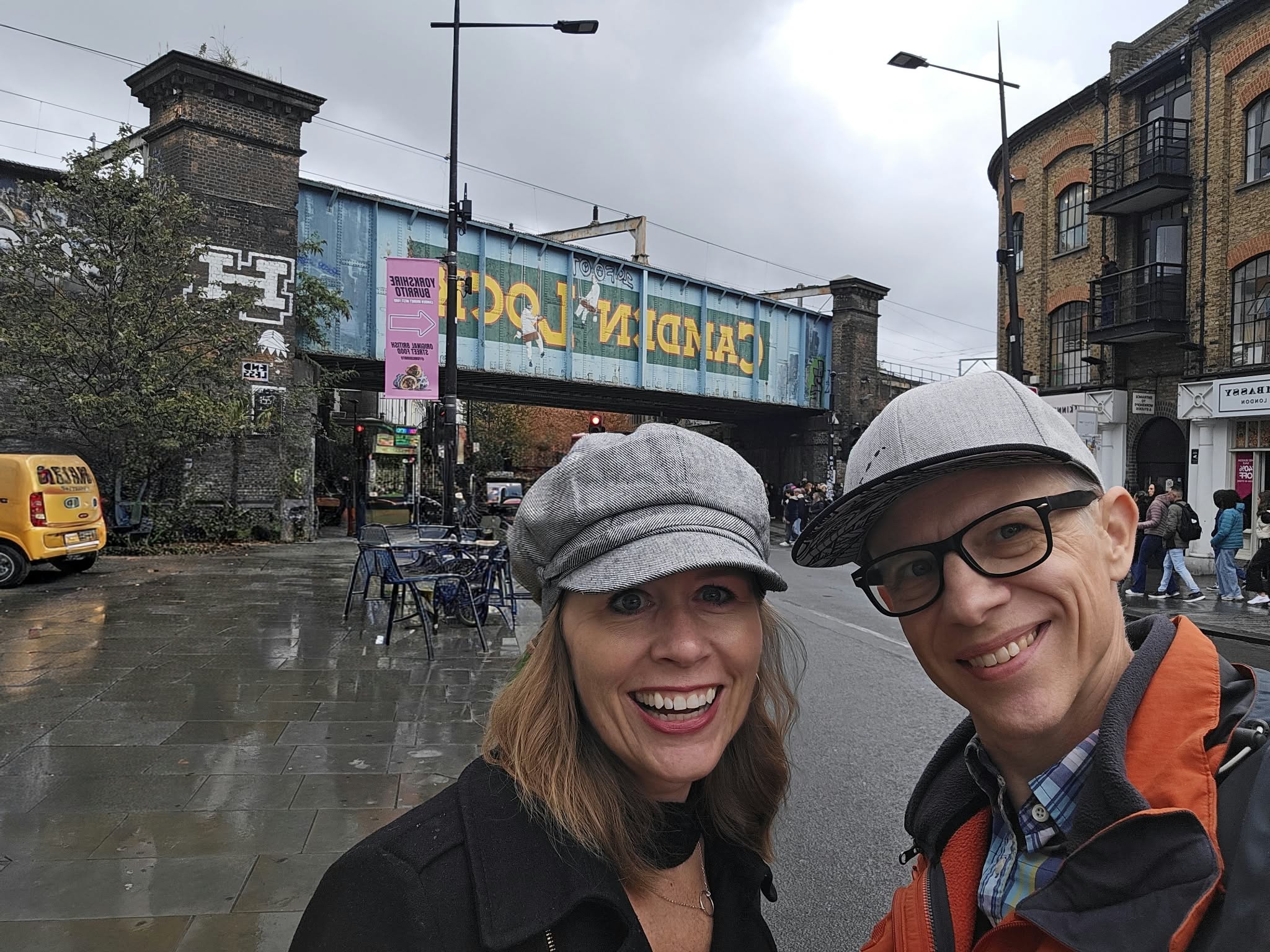 Two smiling women wearing hats and glasses stand outdoors on a rainy day, with a bridge and city buildings in the background.