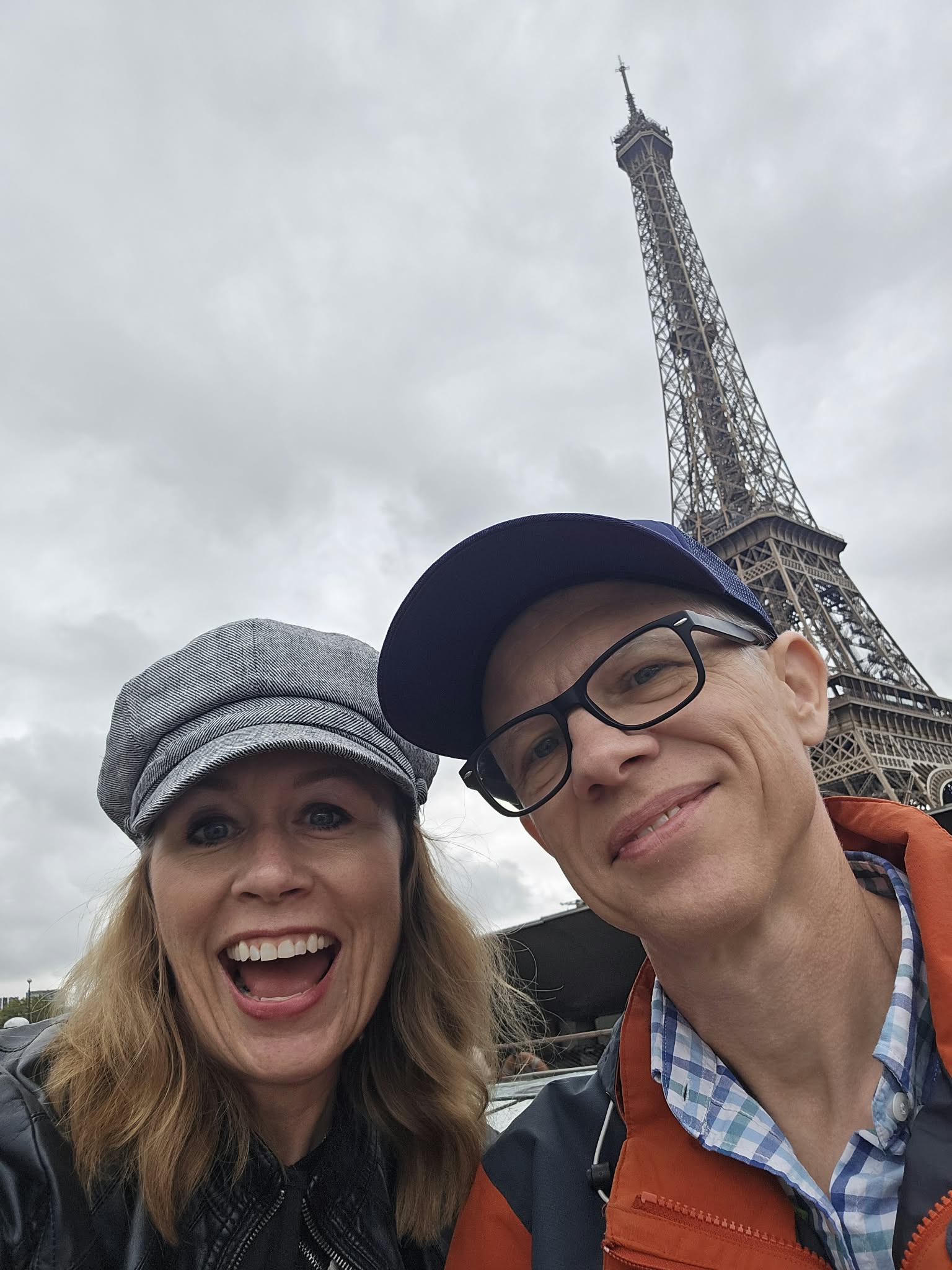 Two people smiling in front of the Eiffel Tower on a cloudy day.