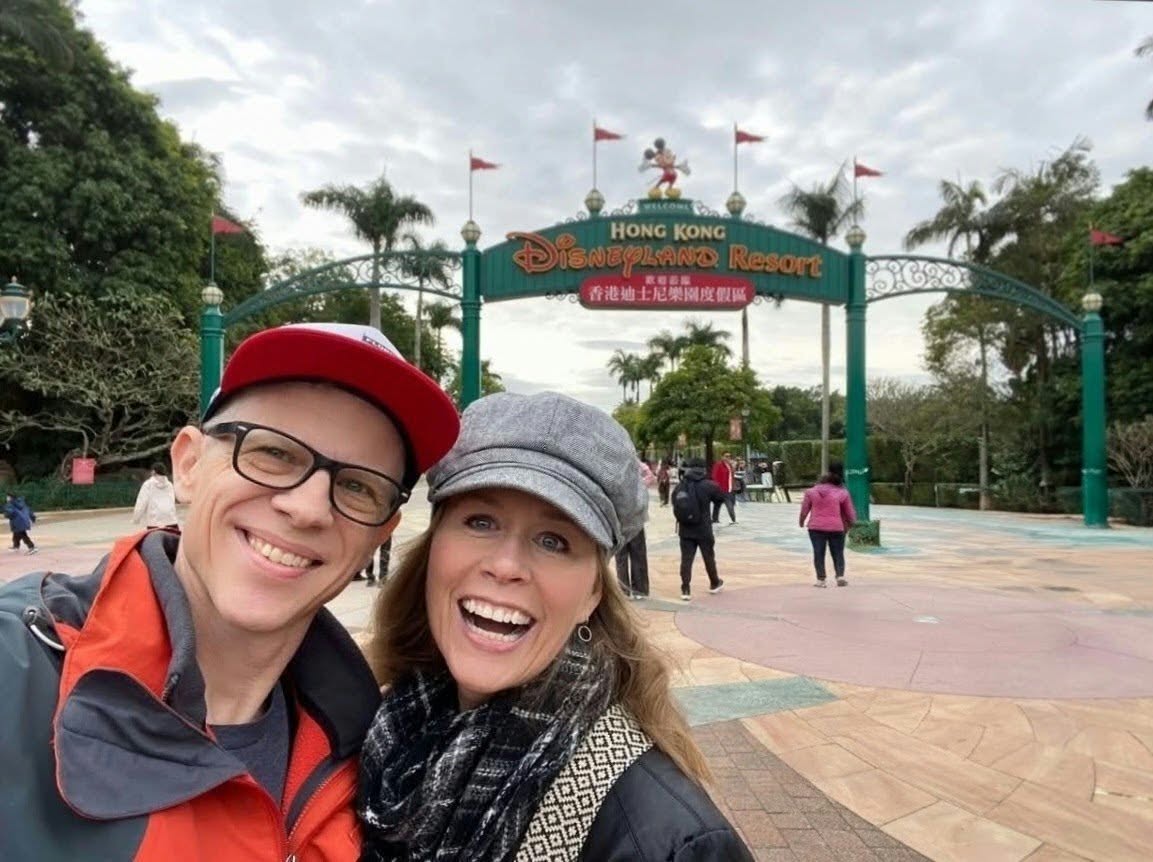 Two smiling people taking a selfie in front of the Hong Kong Disneyland Resort entrance with flags and trees in the background.
