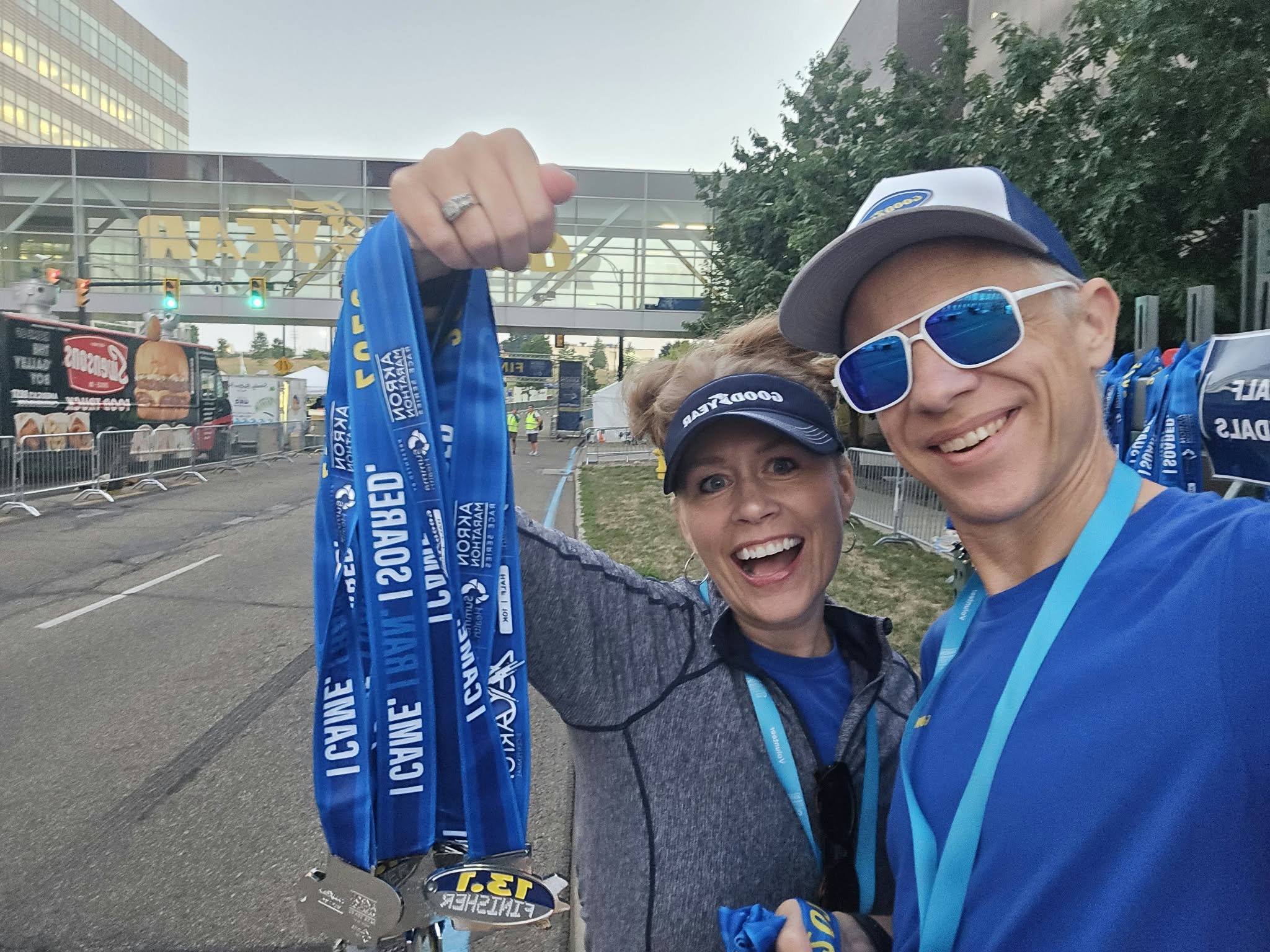 Two smiling people wearing medals and blue clothing, celebrating outdoors with a building and trees in the background.