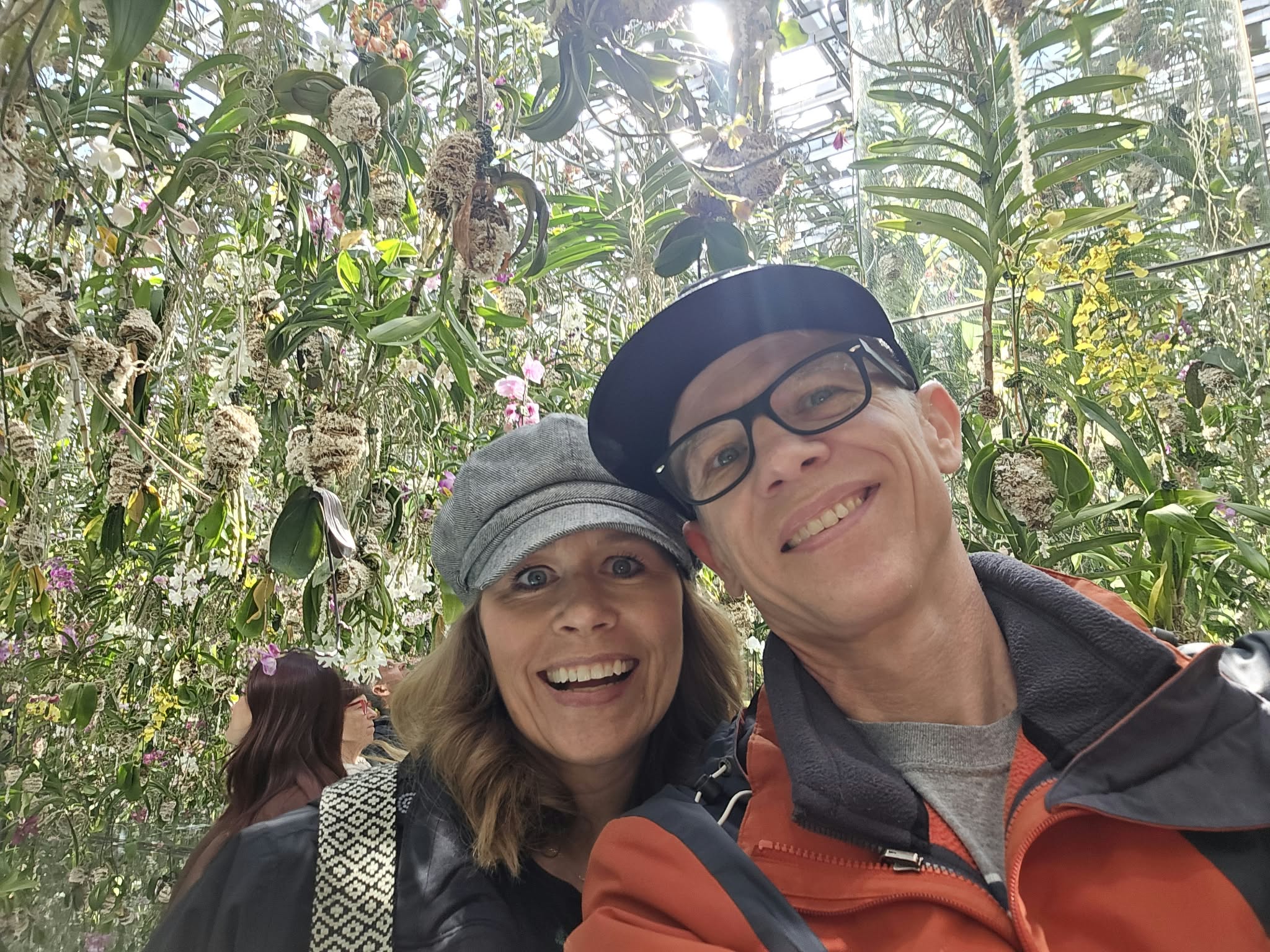Two people smiling outdoors among trees and plants, one wearing a gray hat and glasses, the other wearing a black cap and glasses.