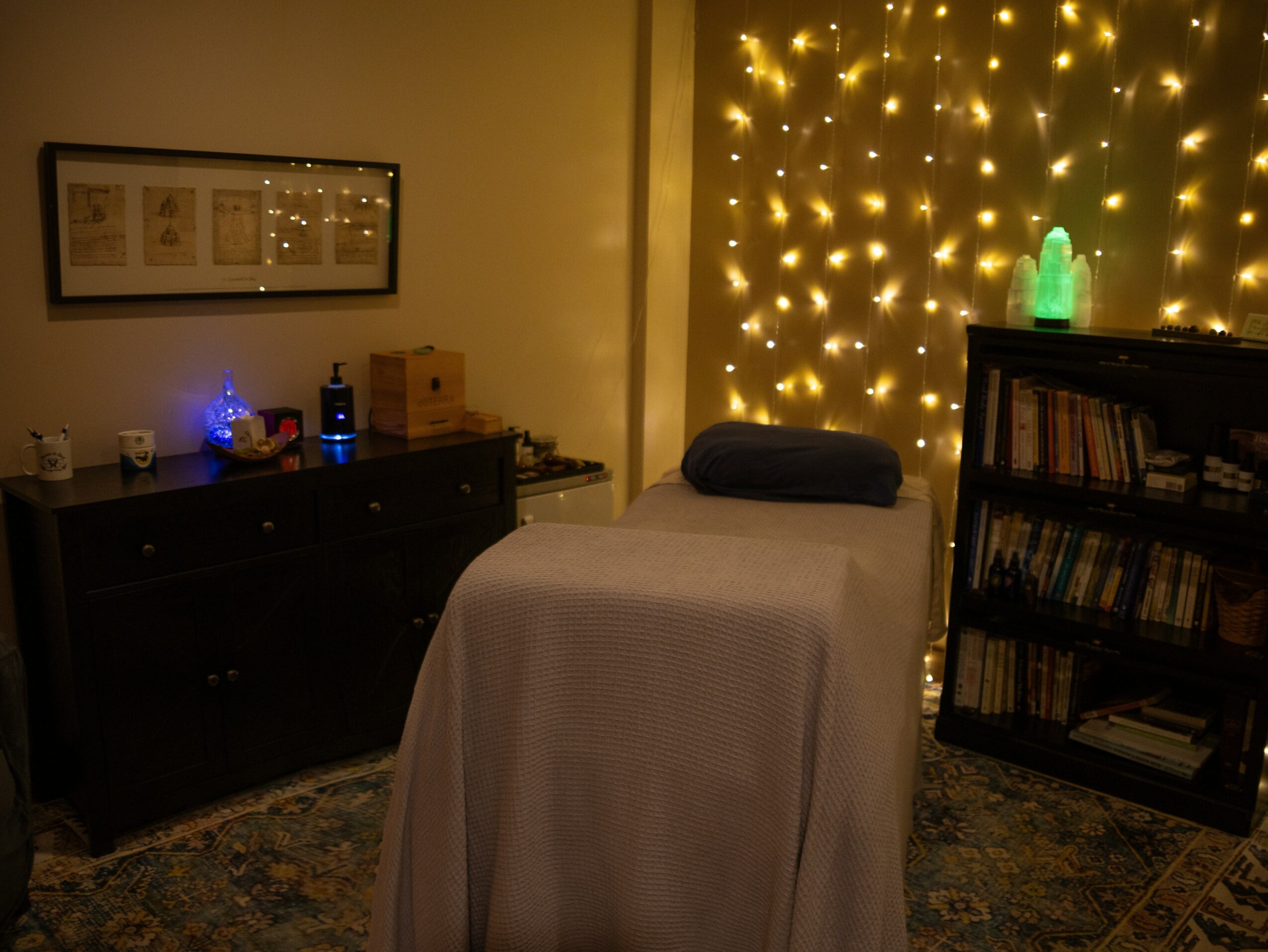 A massage table with a dark pillow in a cozy room decorated with string lights and a bookshelf.